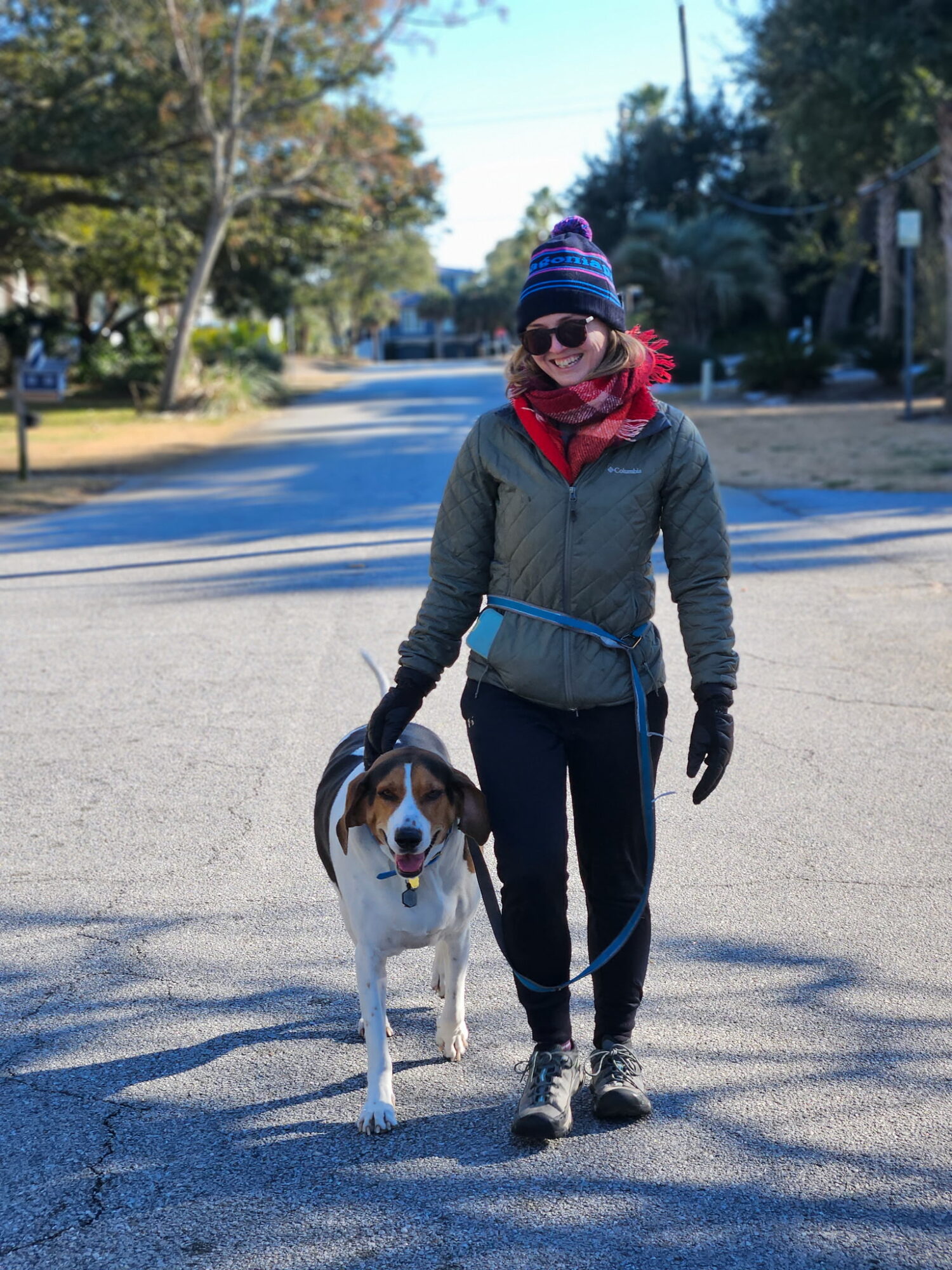 Person walking a dog on a street with trees and houses in the background, wearing sunglasses, a hat, and a jacket.