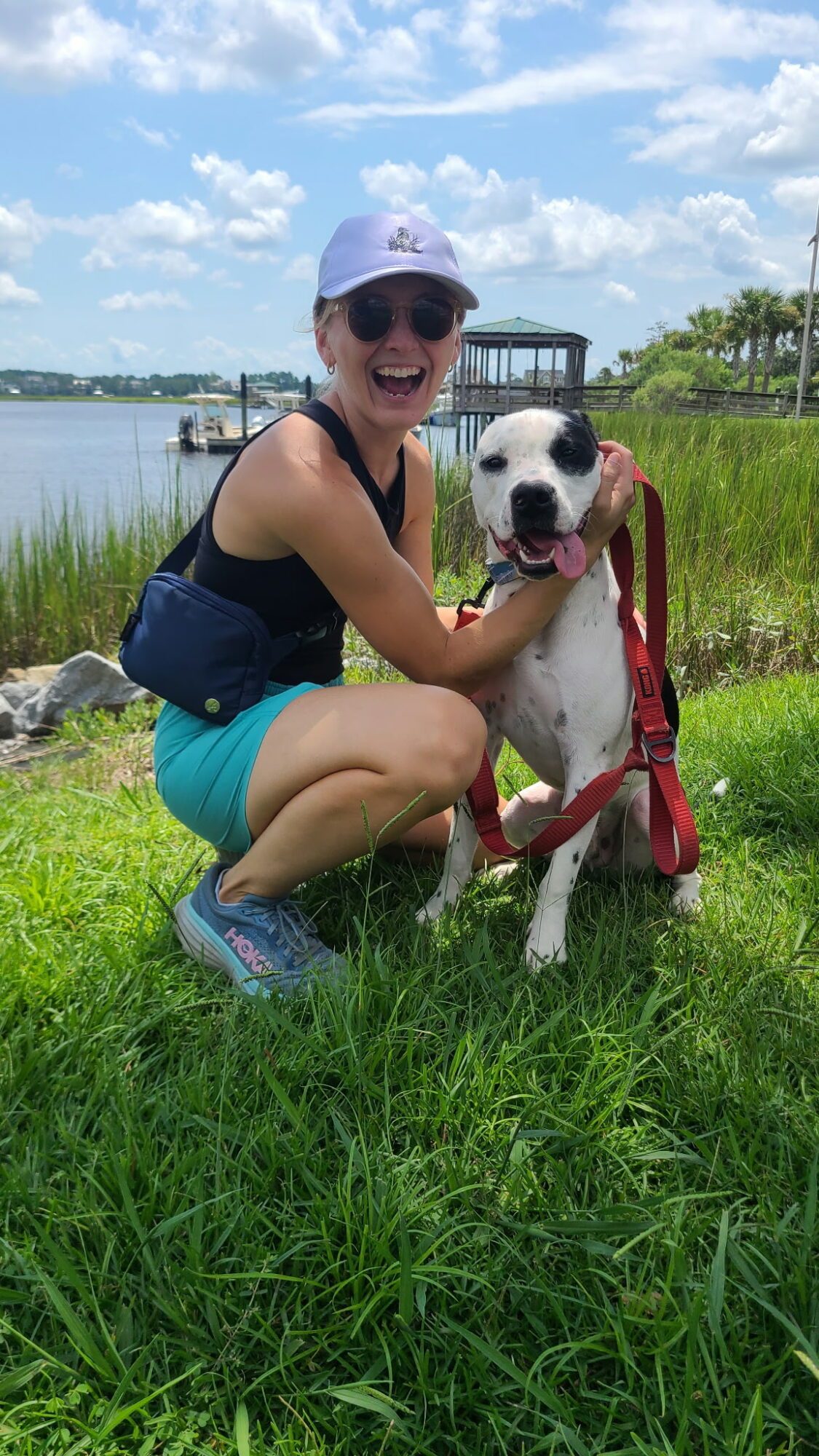 Woman crouching on grass with a dog near water, smiling, wearing sunglasses and a hat.
