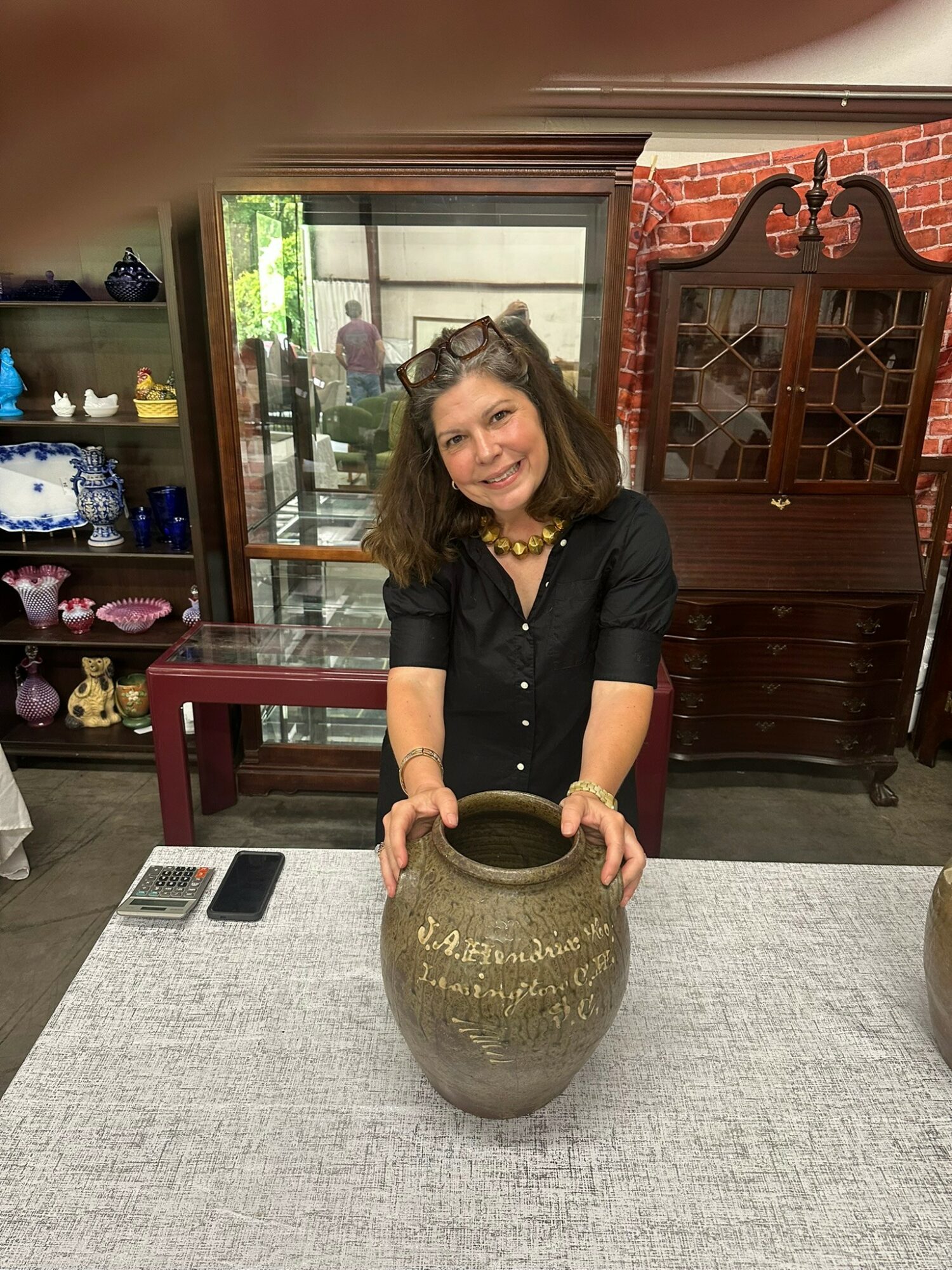 Woman with curly hair and black shirt smiling, holding a large ceramic vase on a table.
