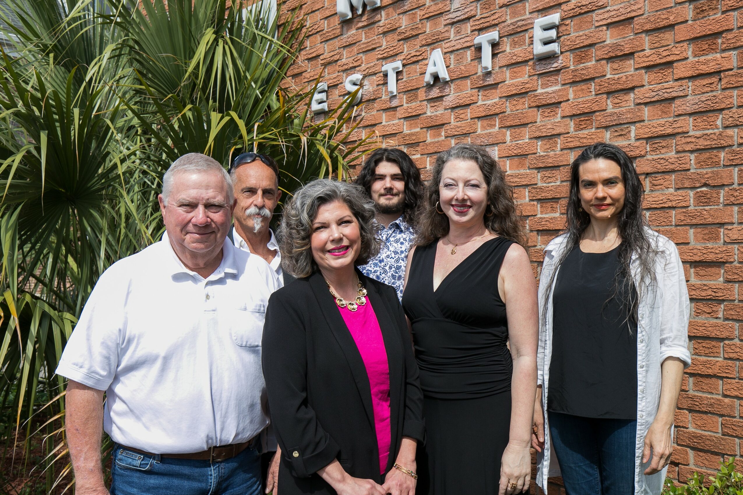 Six people standing outdoors in front of a brick wall with plants, smiling for the photo.