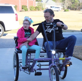 Man riding a purple tricycle with a young girl sitting in the back seat, both outdoors on a sunny day.