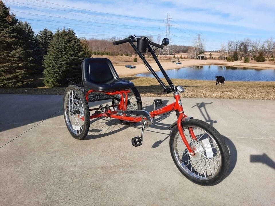 Red tricycle with black seat and large rear wheels parked on concrete near a pond and trees.