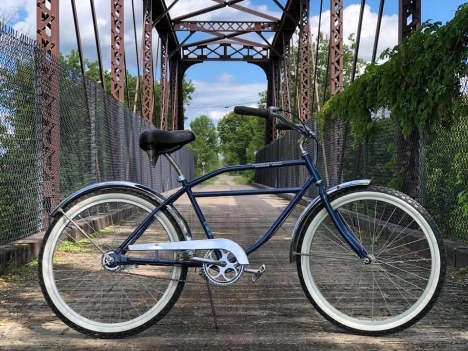 Blue bicycle with black seat and white tires parked on a wooden bridge with metal structure and green trees in background.