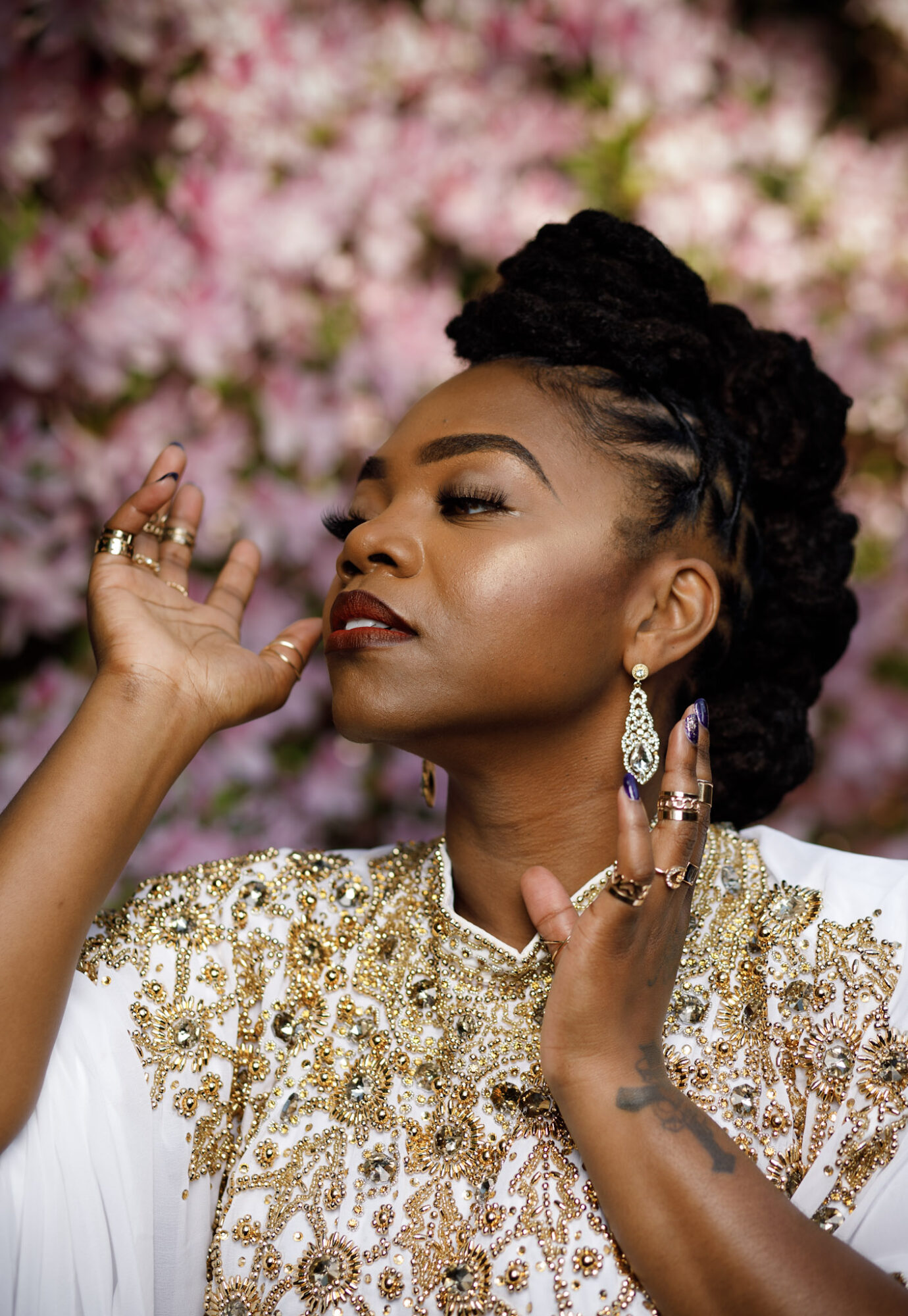 Woman with styled hair and jewelry, posing against pink flowers background.