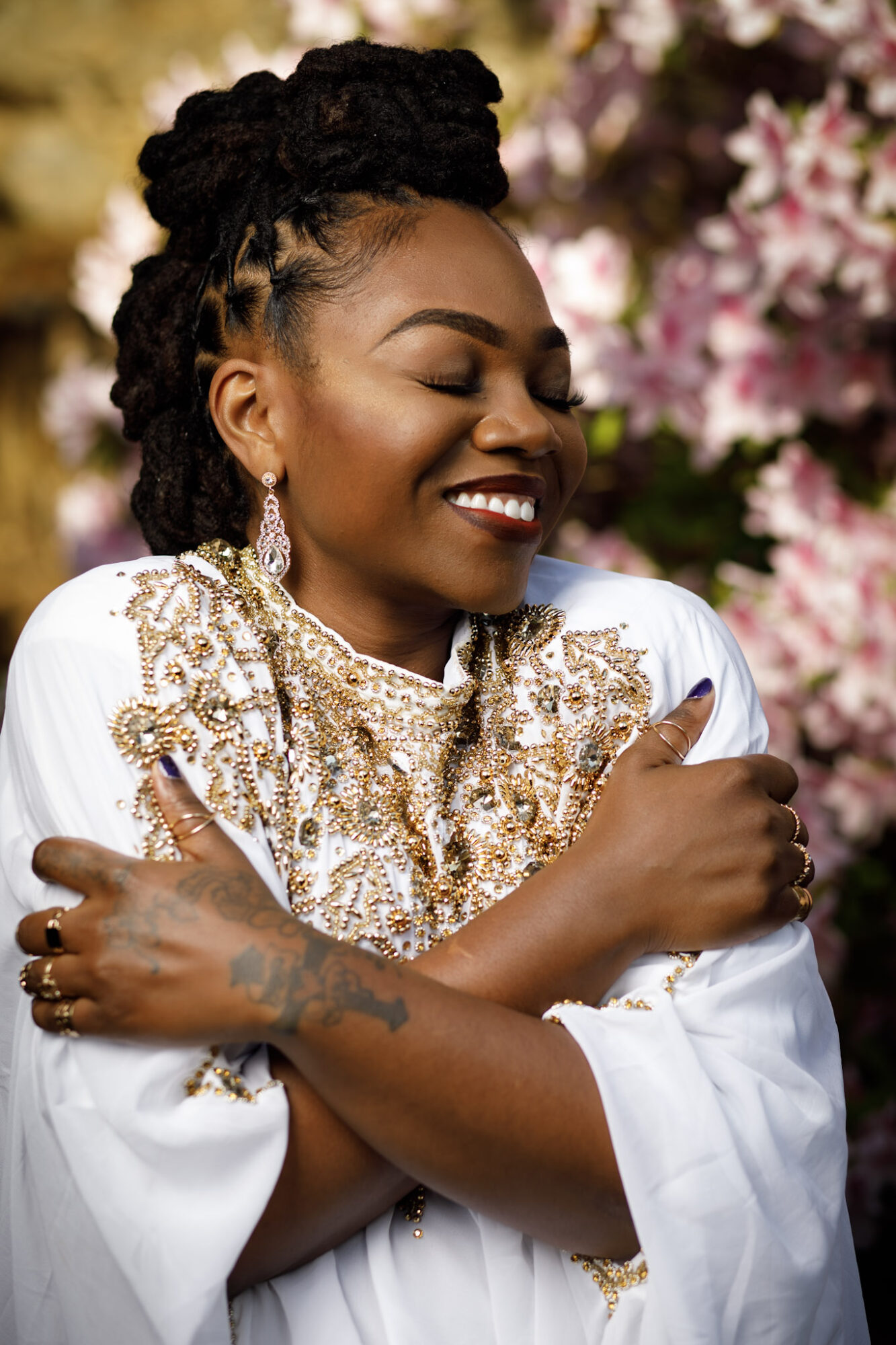 Woman with dreadlocks smiling, wearing a white and gold embroidered outfit, surrounded by pink flowers.