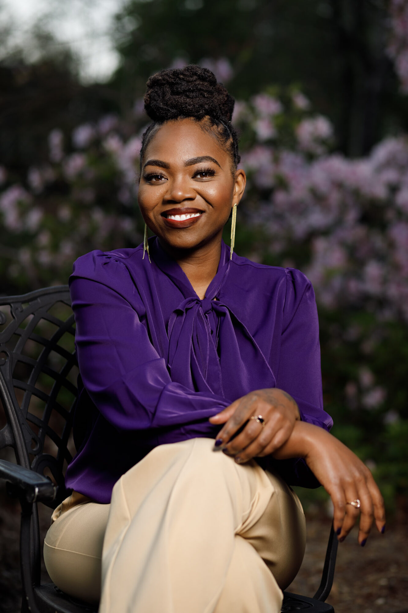 Woman with braided hair smiling outdoors, wearing a purple blouse and beige pants, sitting on a chair.