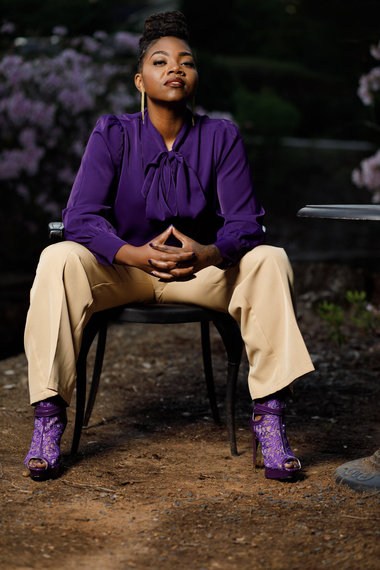 Woman sitting on a chair outdoors with purple flowers in background, wearing purple top, beige pants, and purple boots.