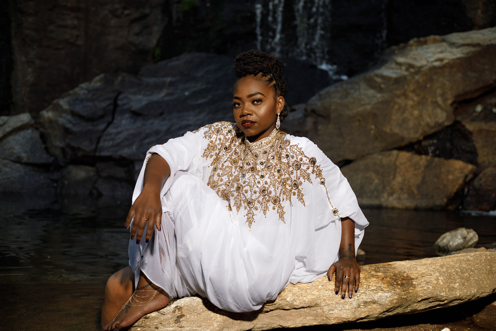 Woman in white dress with gold embellishments sitting on a rock near a waterfall and rocks.