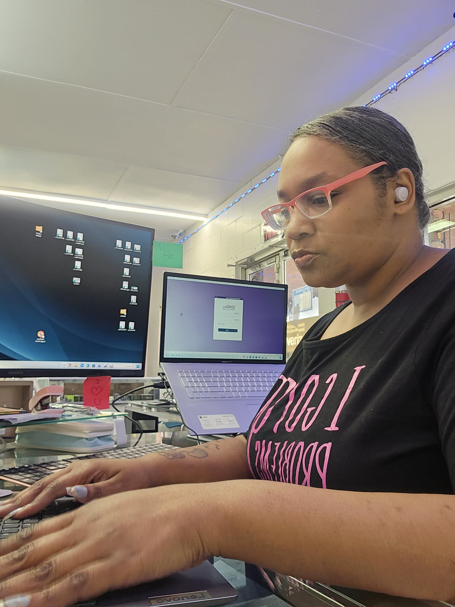 Woman with glasses working at desk with computer and laptop in a room with shelves and books.
