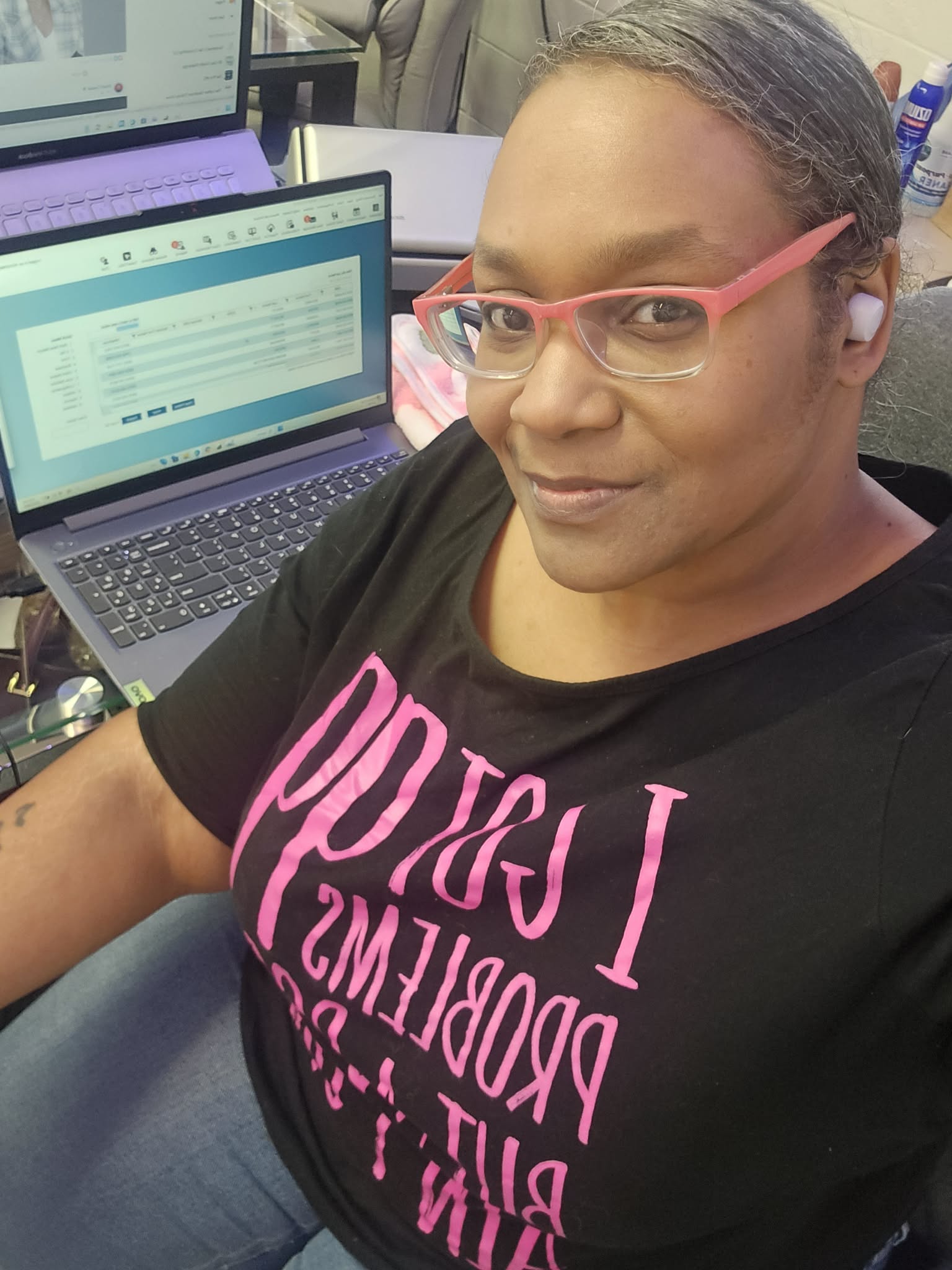 Person with pink glasses taking a selfie in front of a laptop with a spreadsheet open.