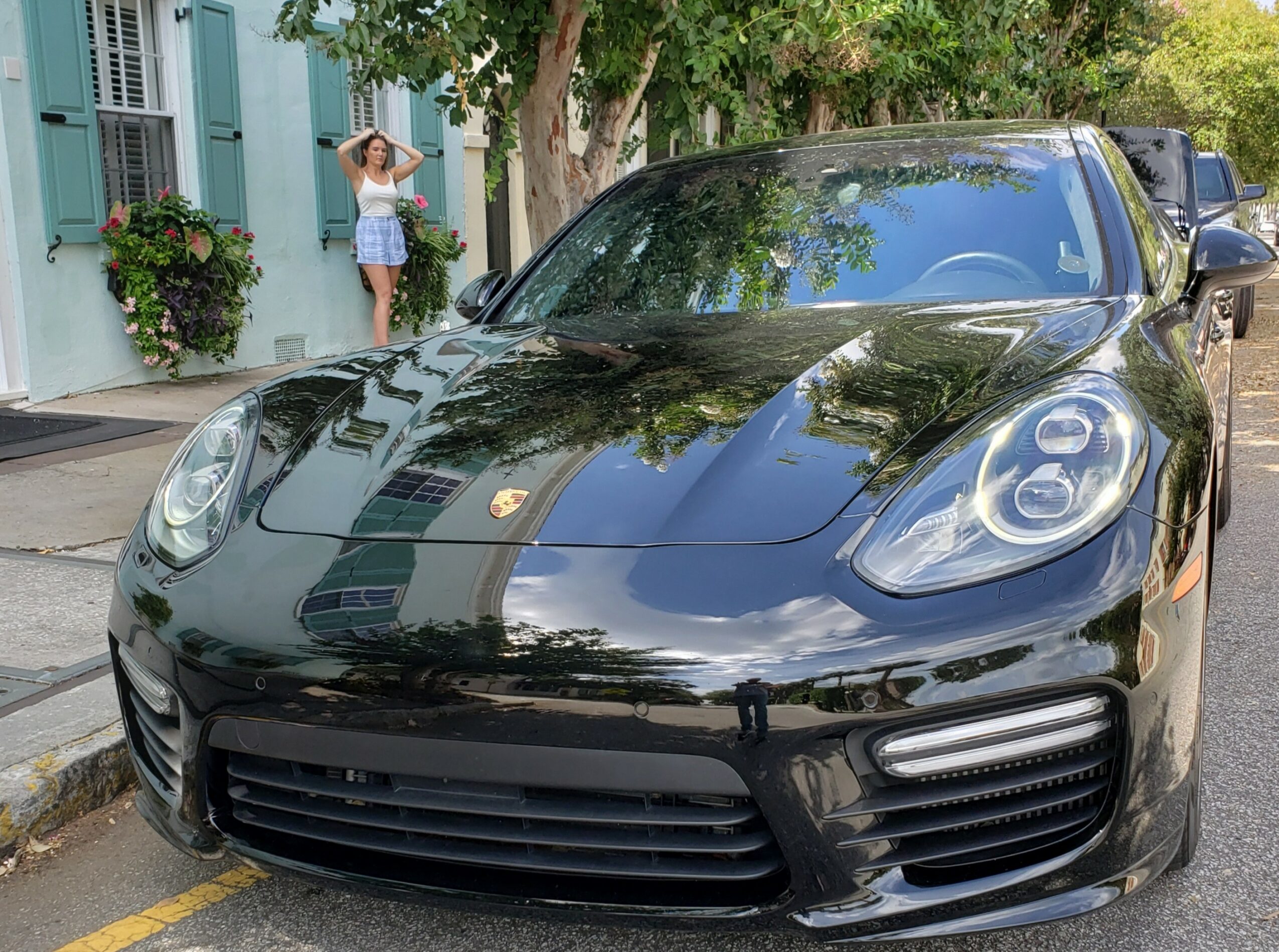 Black sports car parked on street with trees and a woman in background near a building.