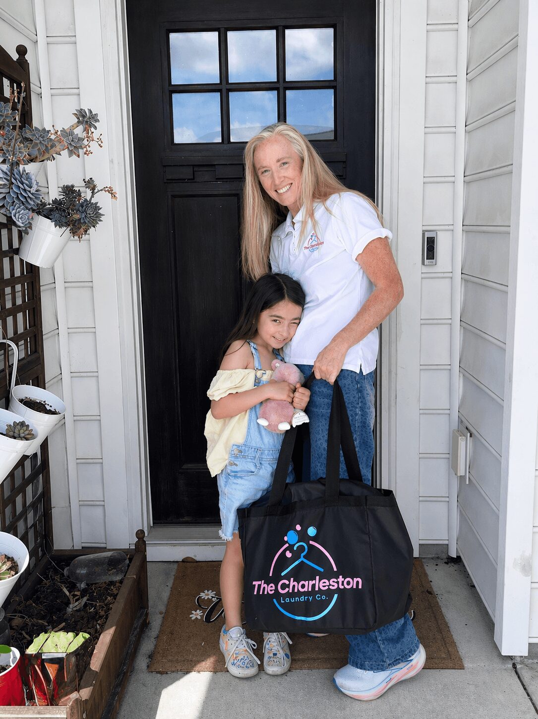 Two females, one adult and one child, standing outside a door with a black frame, holding a black bag with a logo.