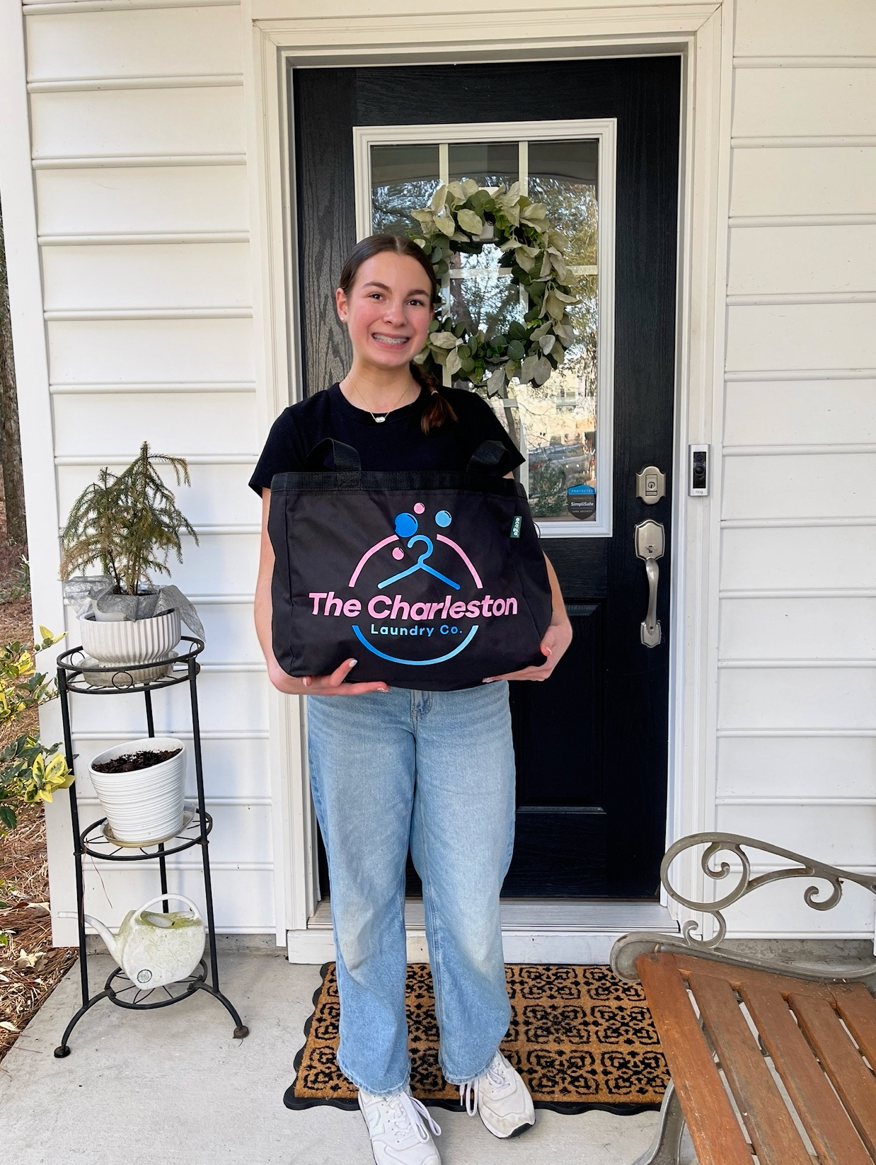 Young woman holding a bag with 'The Charleston' logo, standing outside a house with a black door and white siding.