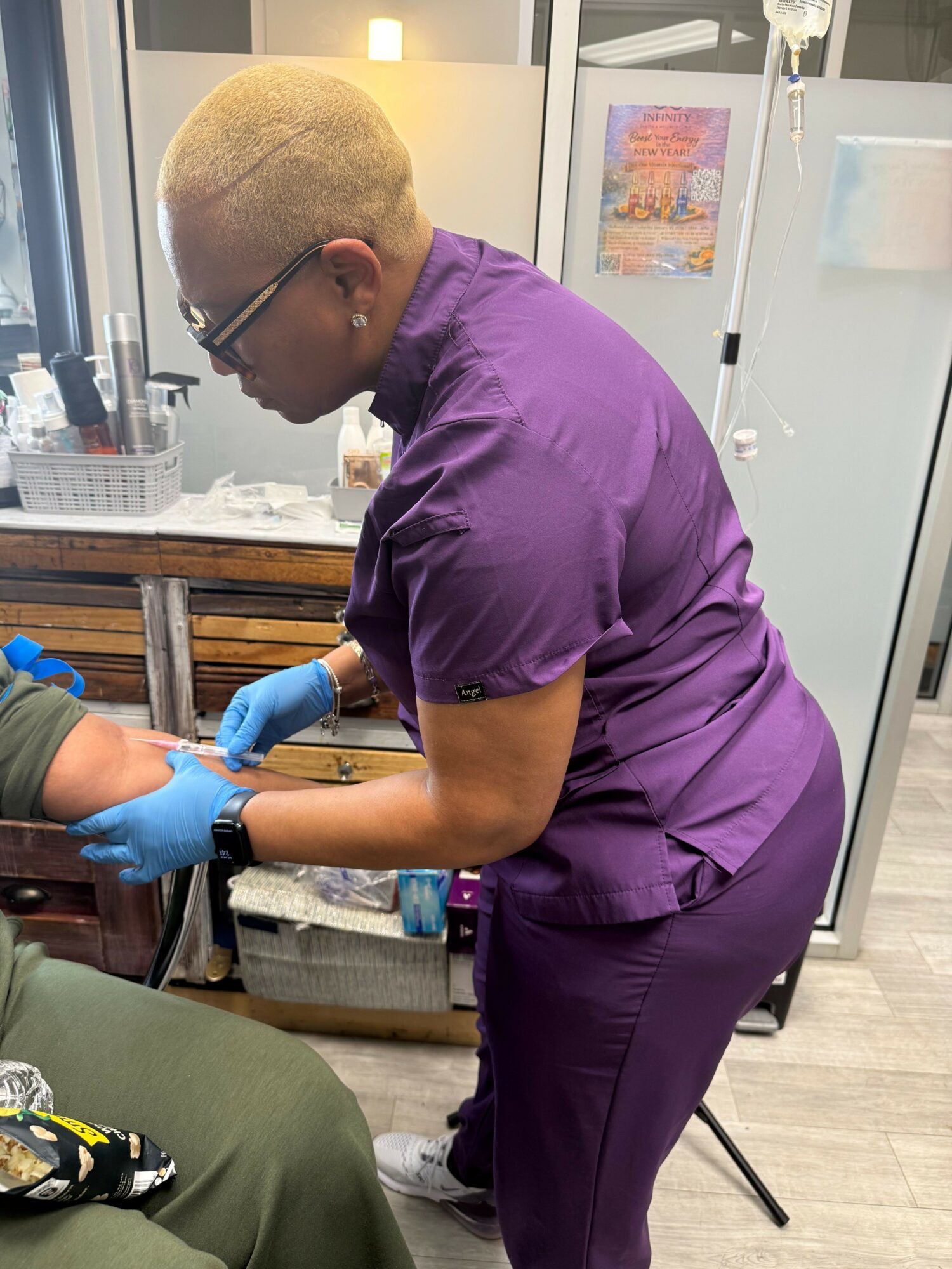 Healthcare worker in purple scrubs and gloves preparing a patient's arm in a clinical setting.