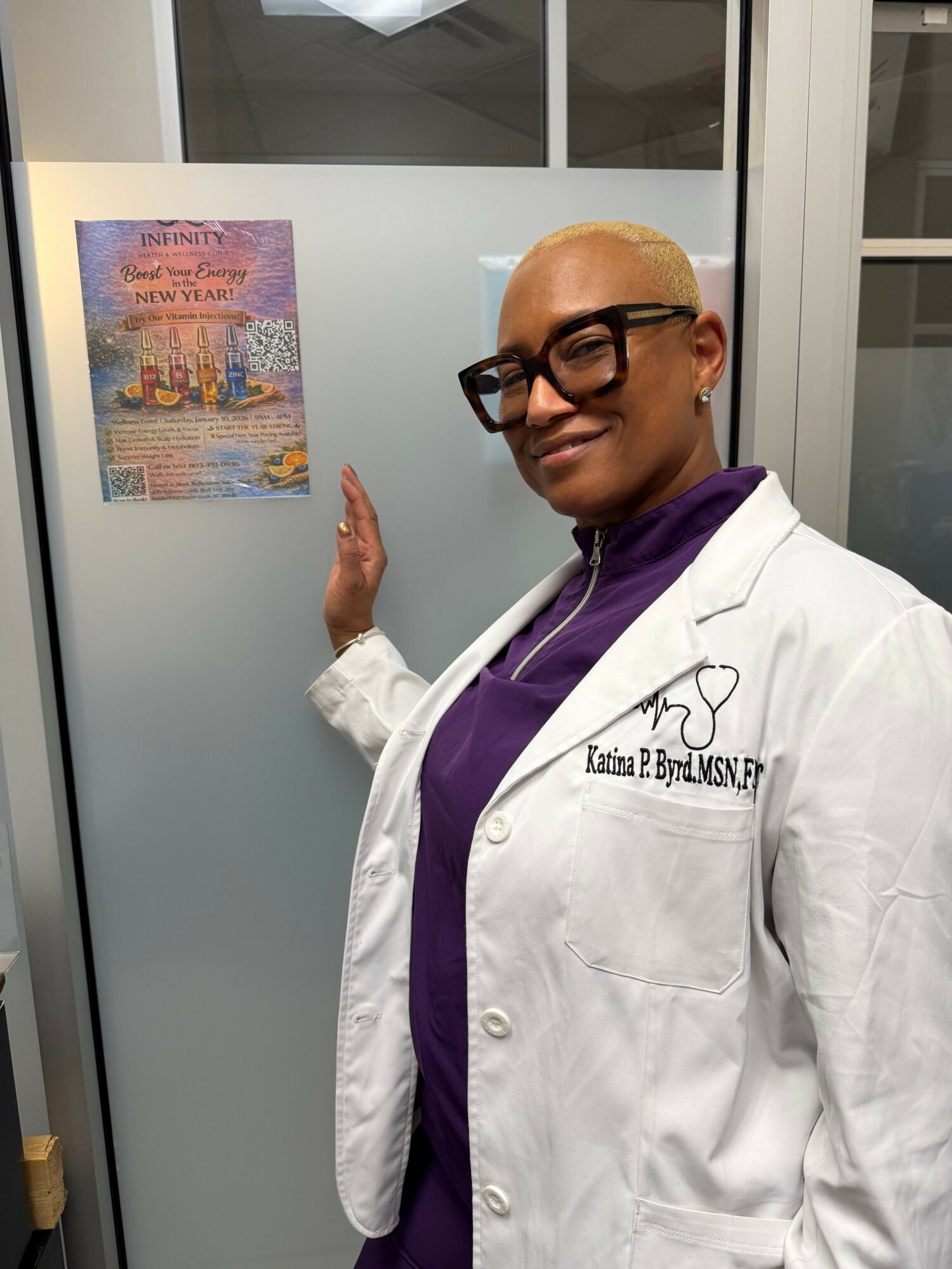 Person with short blonde hair, glasses, white coat, purple shirt, standing next to frosted glass door with poster.