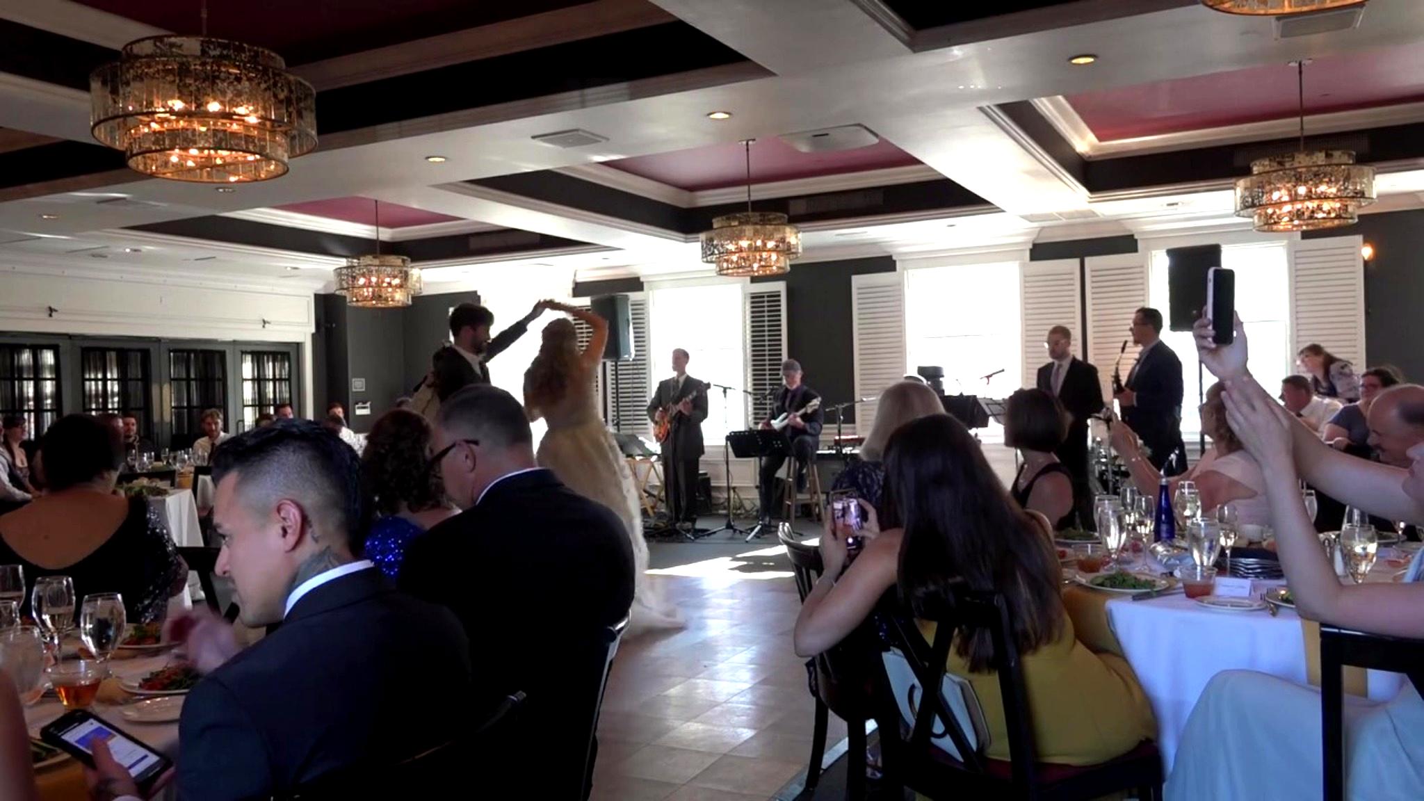 People seated at tables and standing in a decorated banquet hall with chandeliers and a band performing.