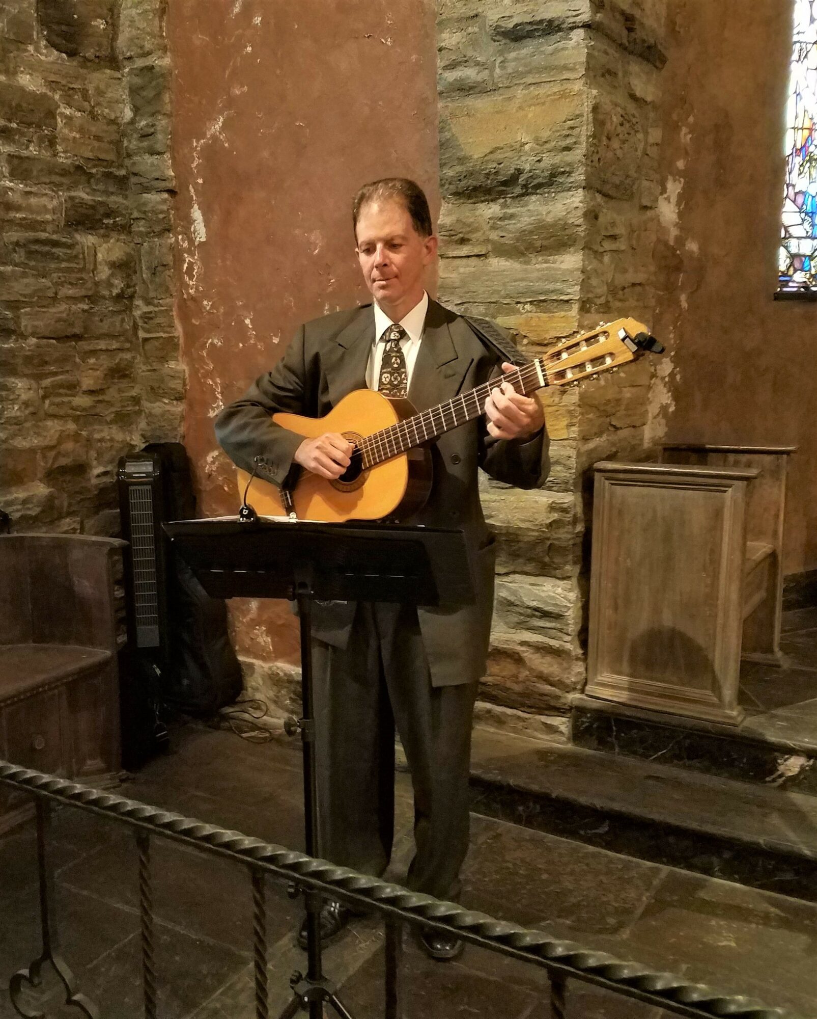 Man in suit playing guitar indoors with stone walls and a music stand.