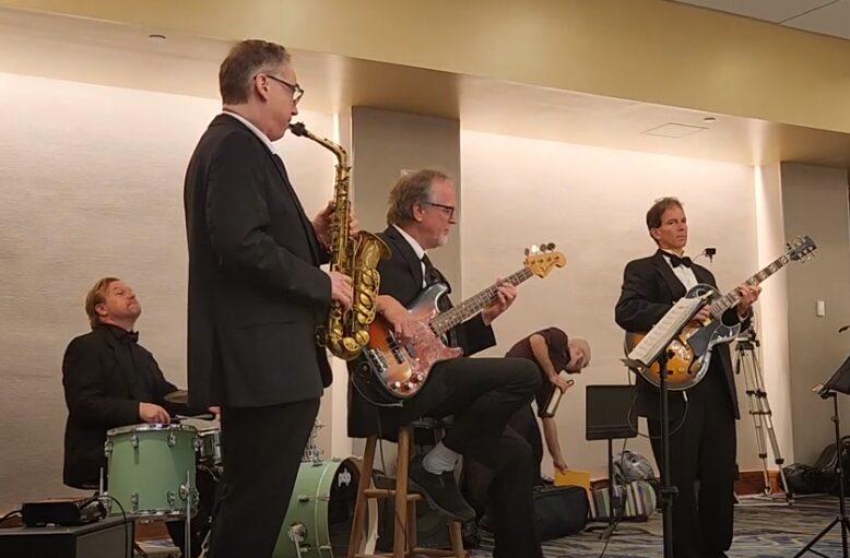 Four musicians performing on stage: a drummer, a saxophonist, and two guitarists, in a concert setting.