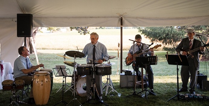 Four musicians perform under a tent outdoors, with instruments including drums, guitar, and percussion, in a grassy area.