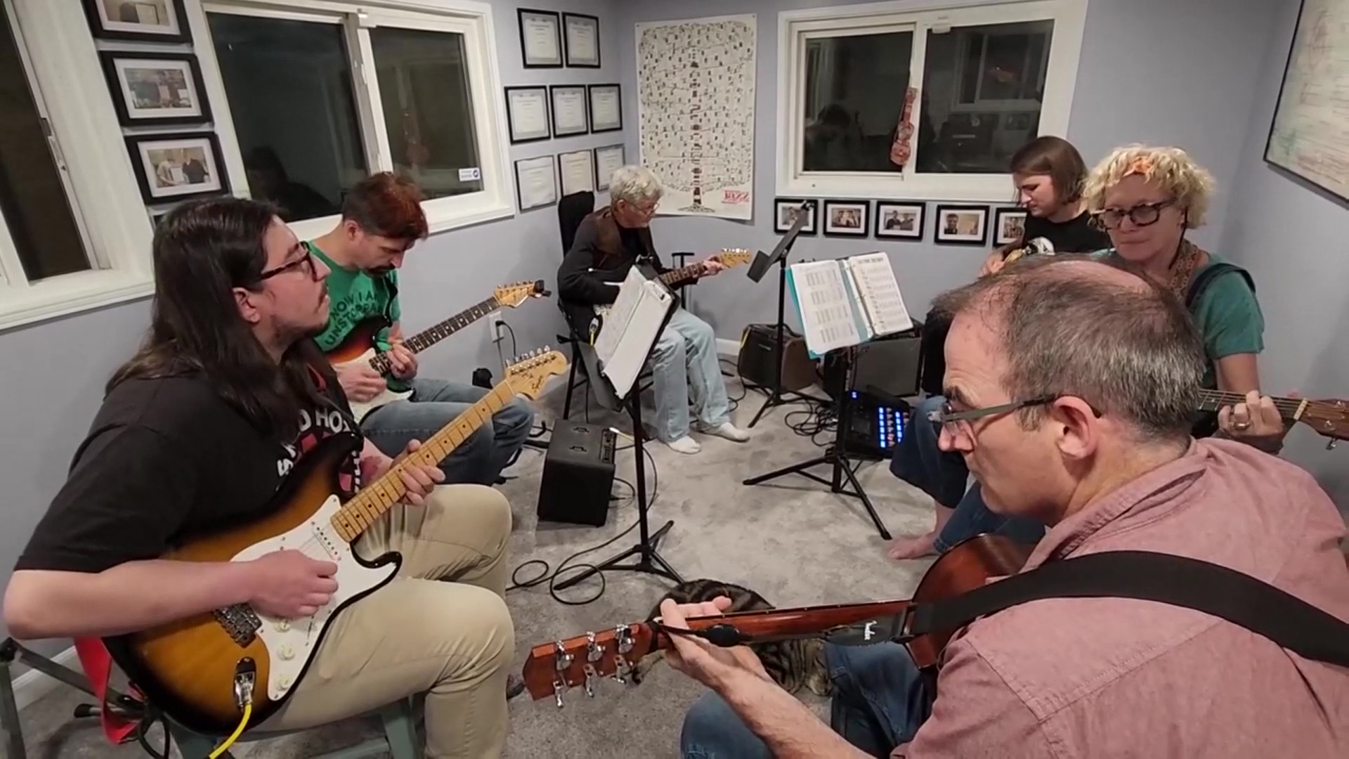 Group of seven people playing guitars and singing in a small room with framed pictures on the walls.