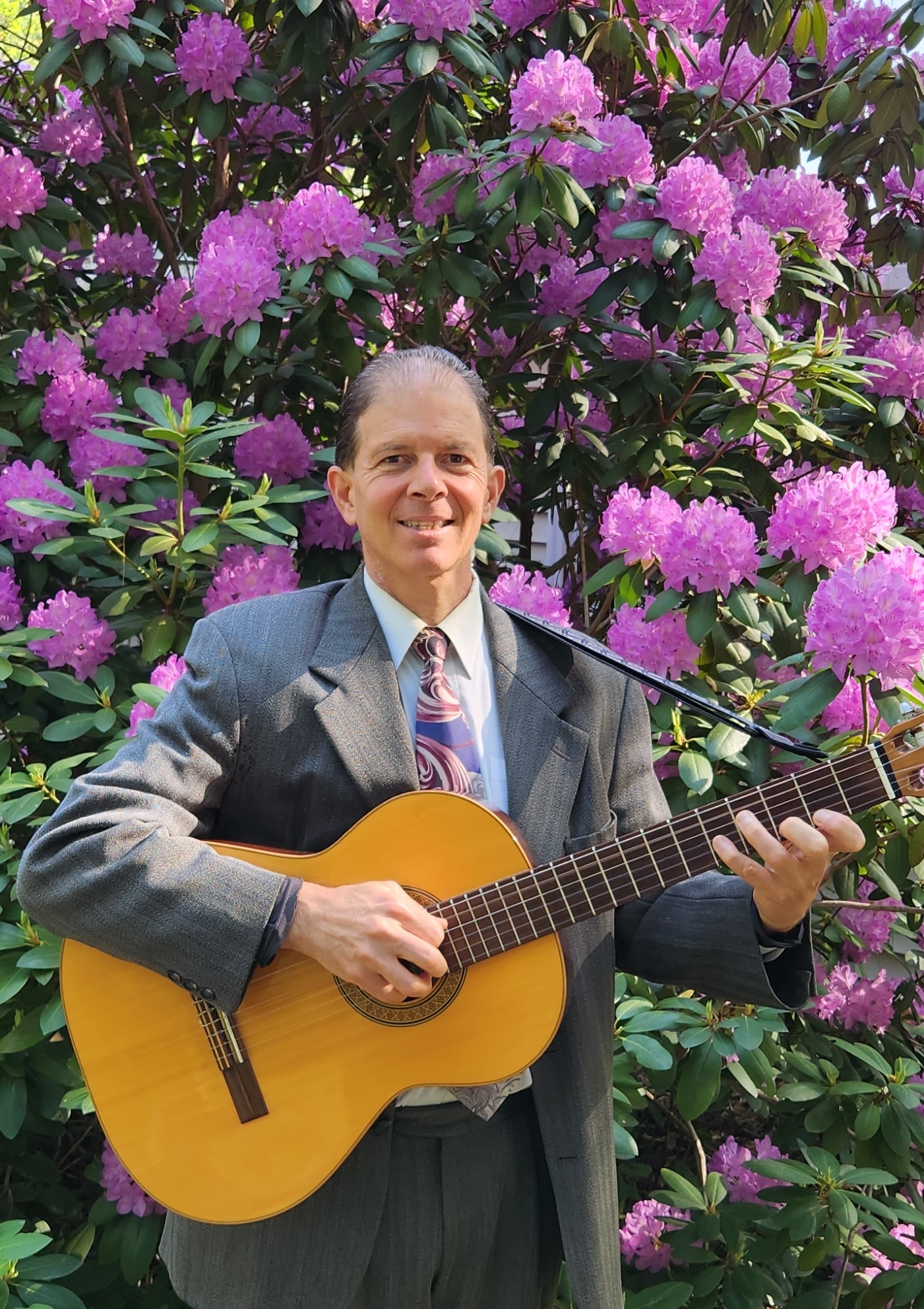 Person in suit holding a guitar in front of blooming purple flowers.