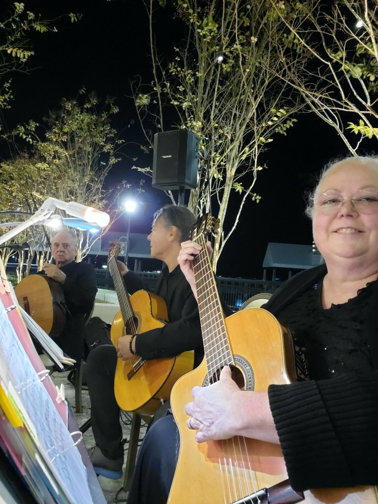 Three people playing guitars outdoors at night, with trees and a speaker in the background.