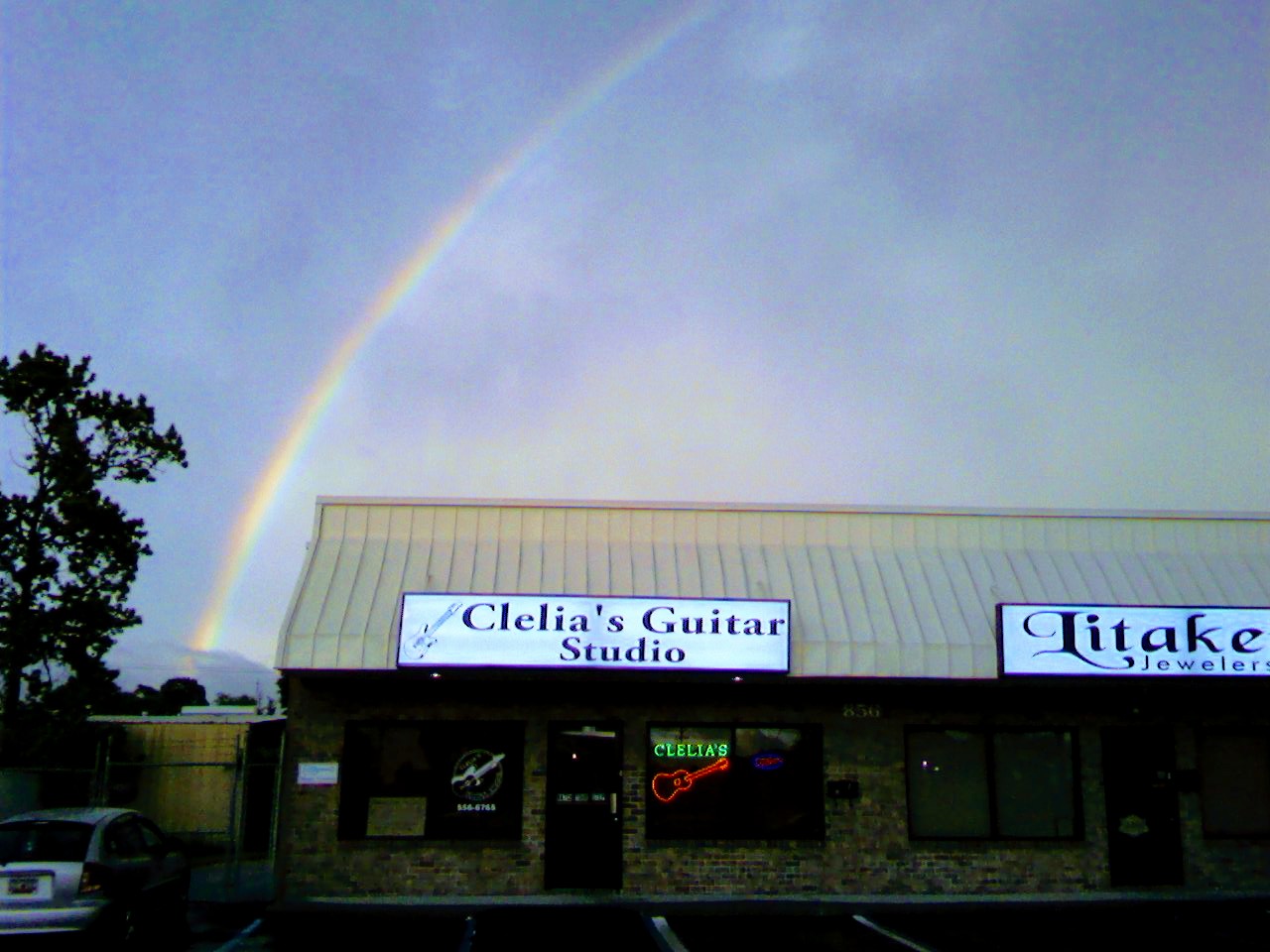 Building with signs for Clelia's Guitar Studio and Litake Jewelry, rainbow in cloudy sky, trees on left side.