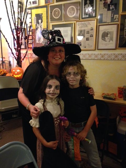 Woman dressed as a witch with two children in a decorated room, Halloween decorations visible.