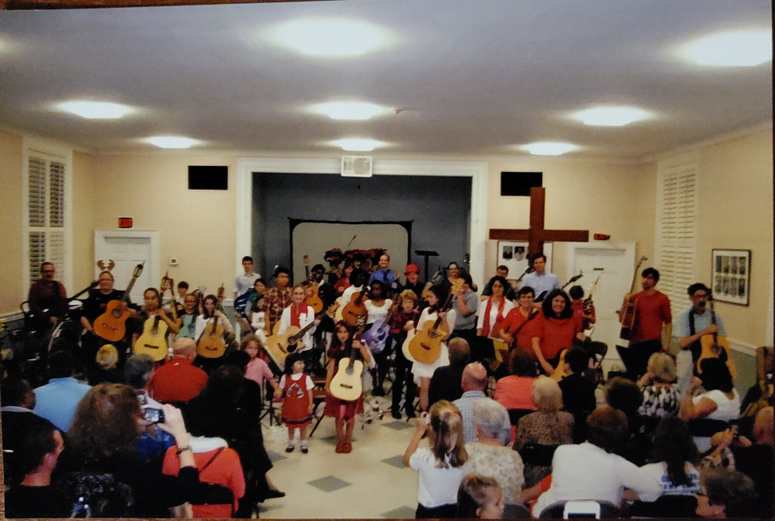 Group of children and adults performing with guitars on stage in a room with audience seated.