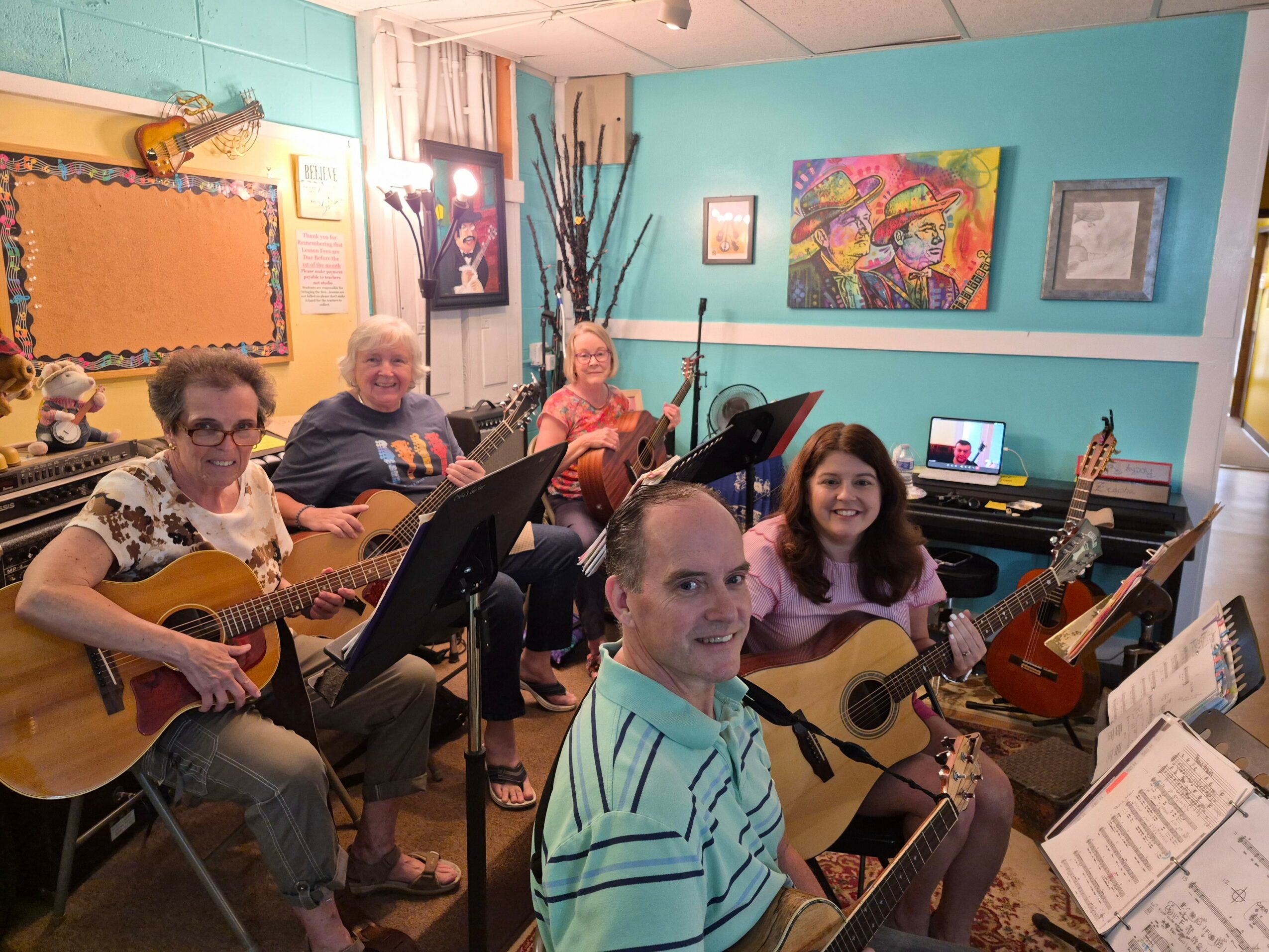 Five people in a music room with guitars, music stands, and colorful wall art, smiling and looking at the camera.