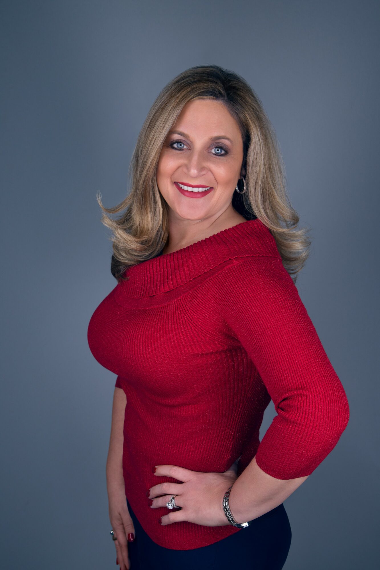 Smiling woman with shoulder-length blonde hair wearing a red top and dark pants, posing with hand on hip.