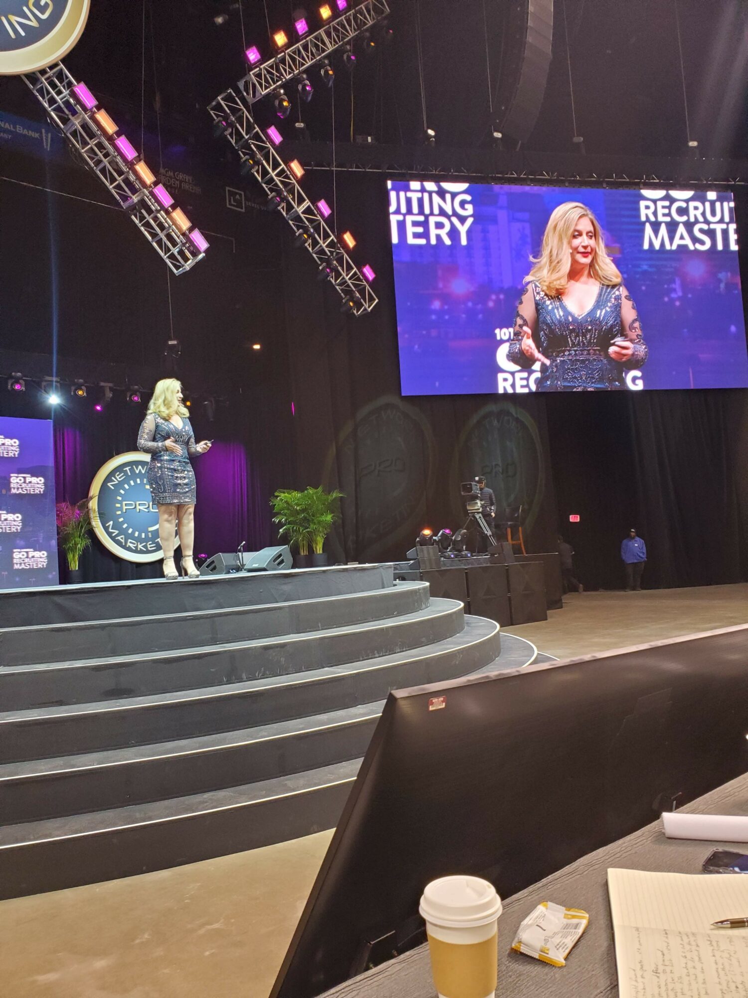 Woman on stage speaking into microphone, large screen behind displaying her image, stage with steps, audience area in foreground.