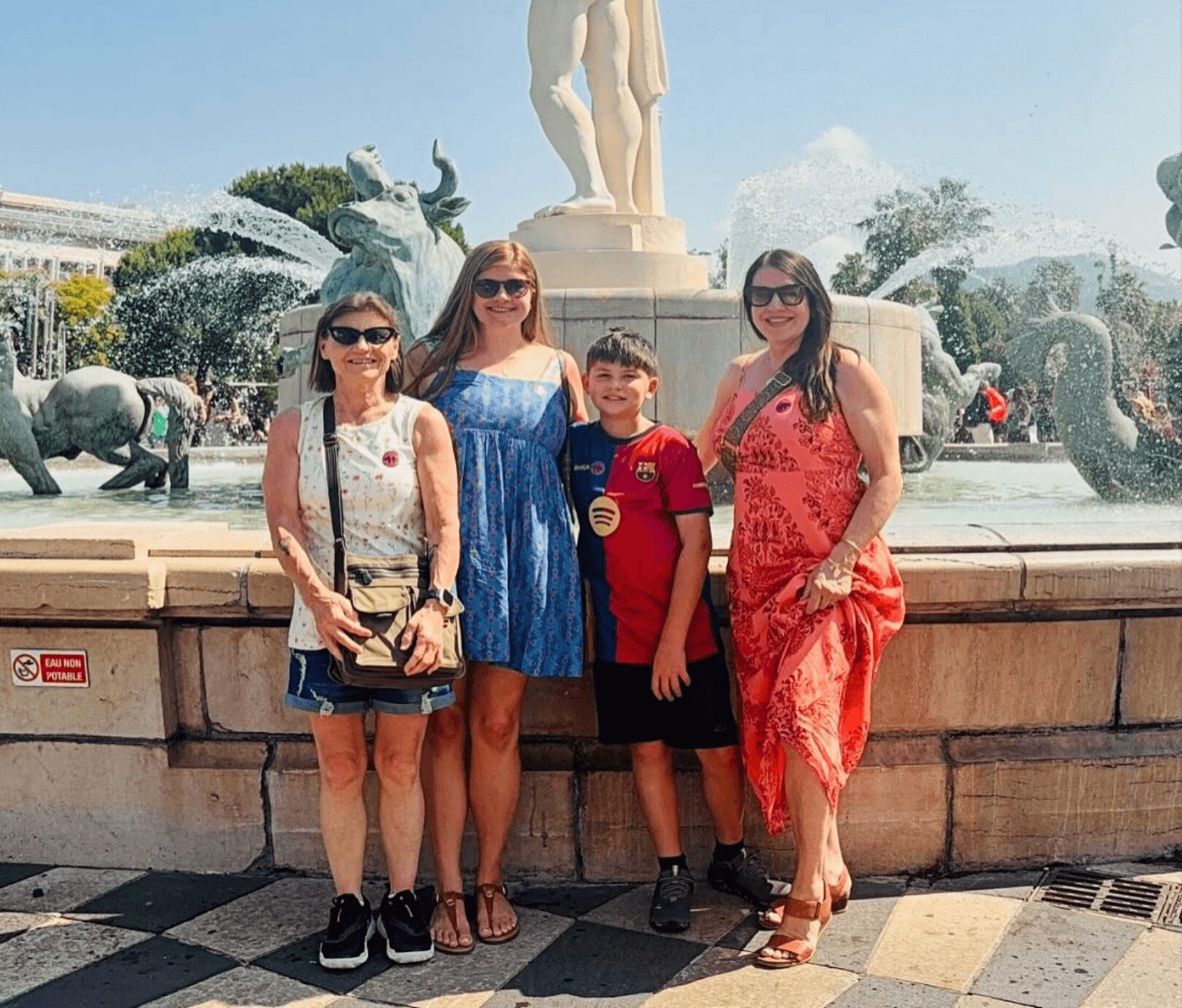 Four people standing in front of a fountain with sculptures, smiling, outdoors on a sunny day.
