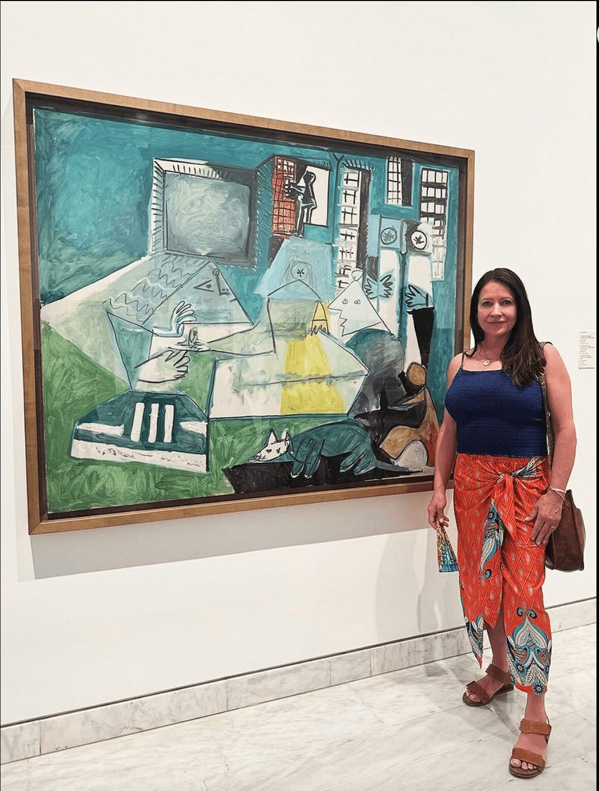 Woman standing next to a colorful abstract painting in a gallery, smiling, with a brown bag on her shoulder.