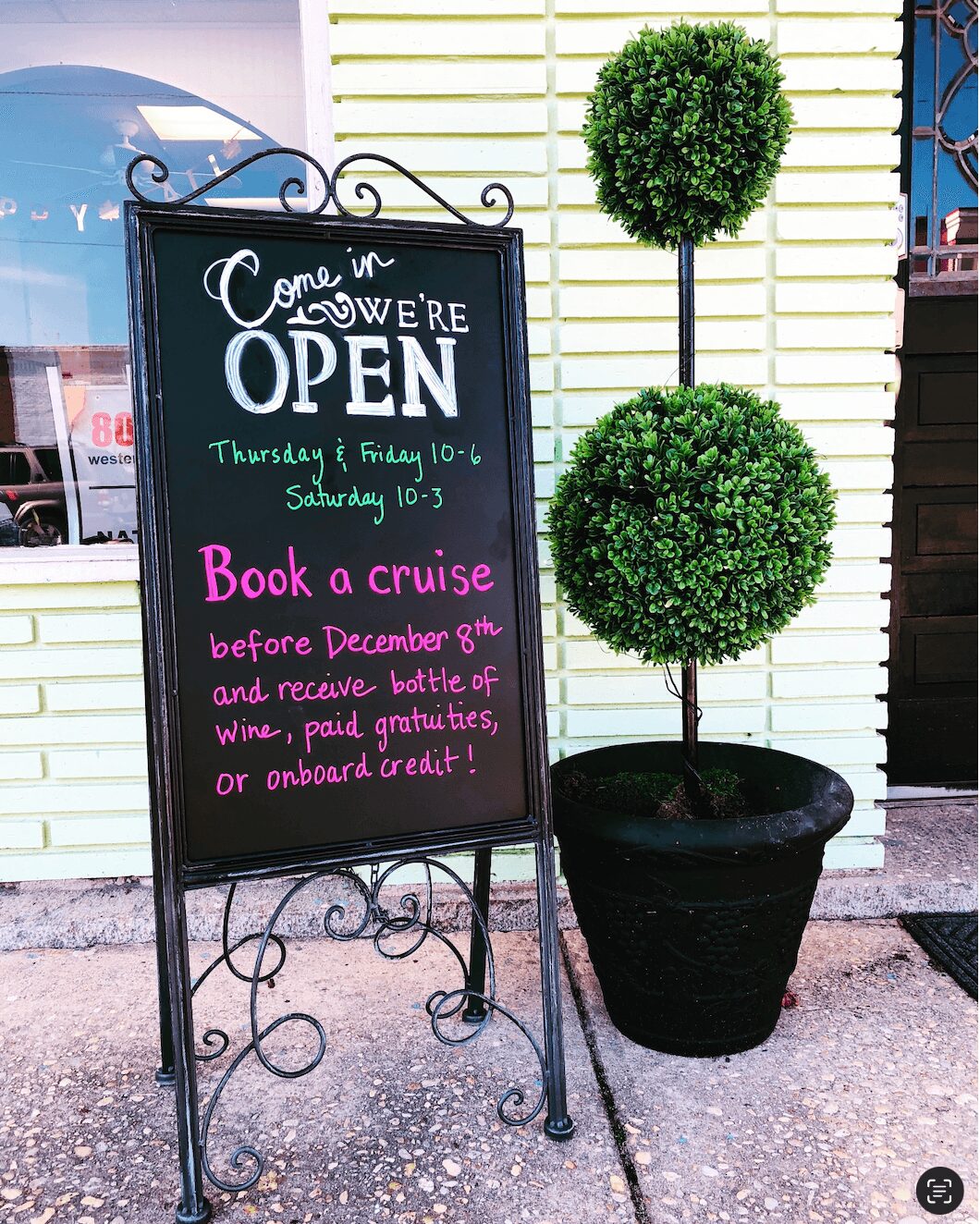 Chalkboard sign with opening hours and promotional message, next to a potted topiary plant outside a building.