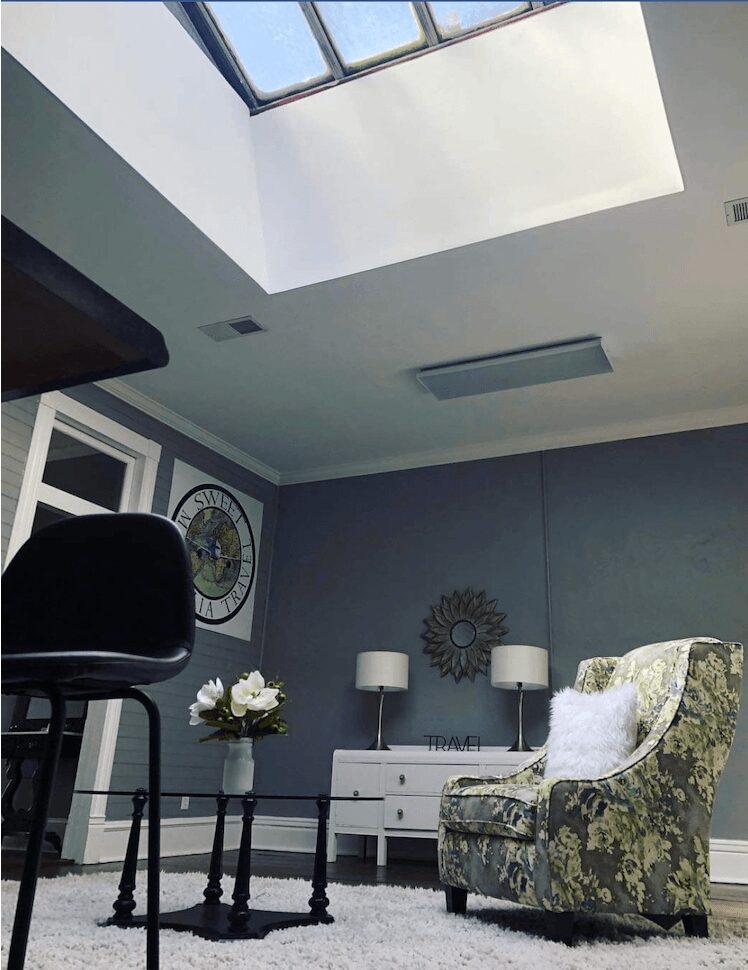 Room with skylight, gray walls, armchair, side table with flowers, and two lamps on a white dresser.