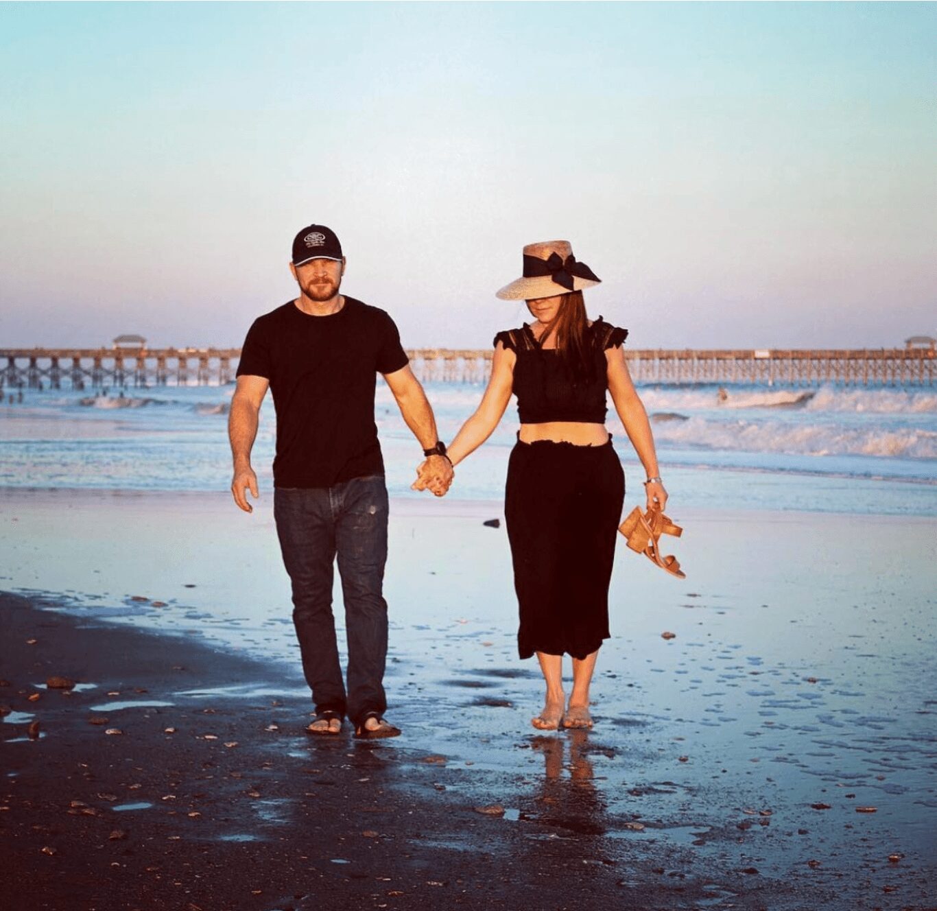A man and woman walk hand in hand along the beach with the ocean and pier in the background.