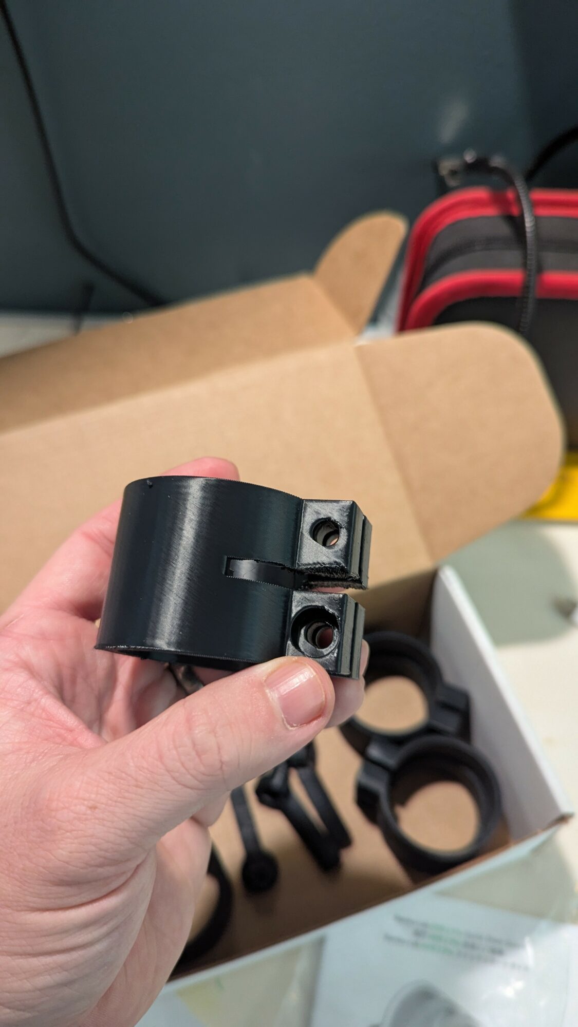 Hand holding a black cylindrical object with two screws, with a cardboard box and various black components in the background.