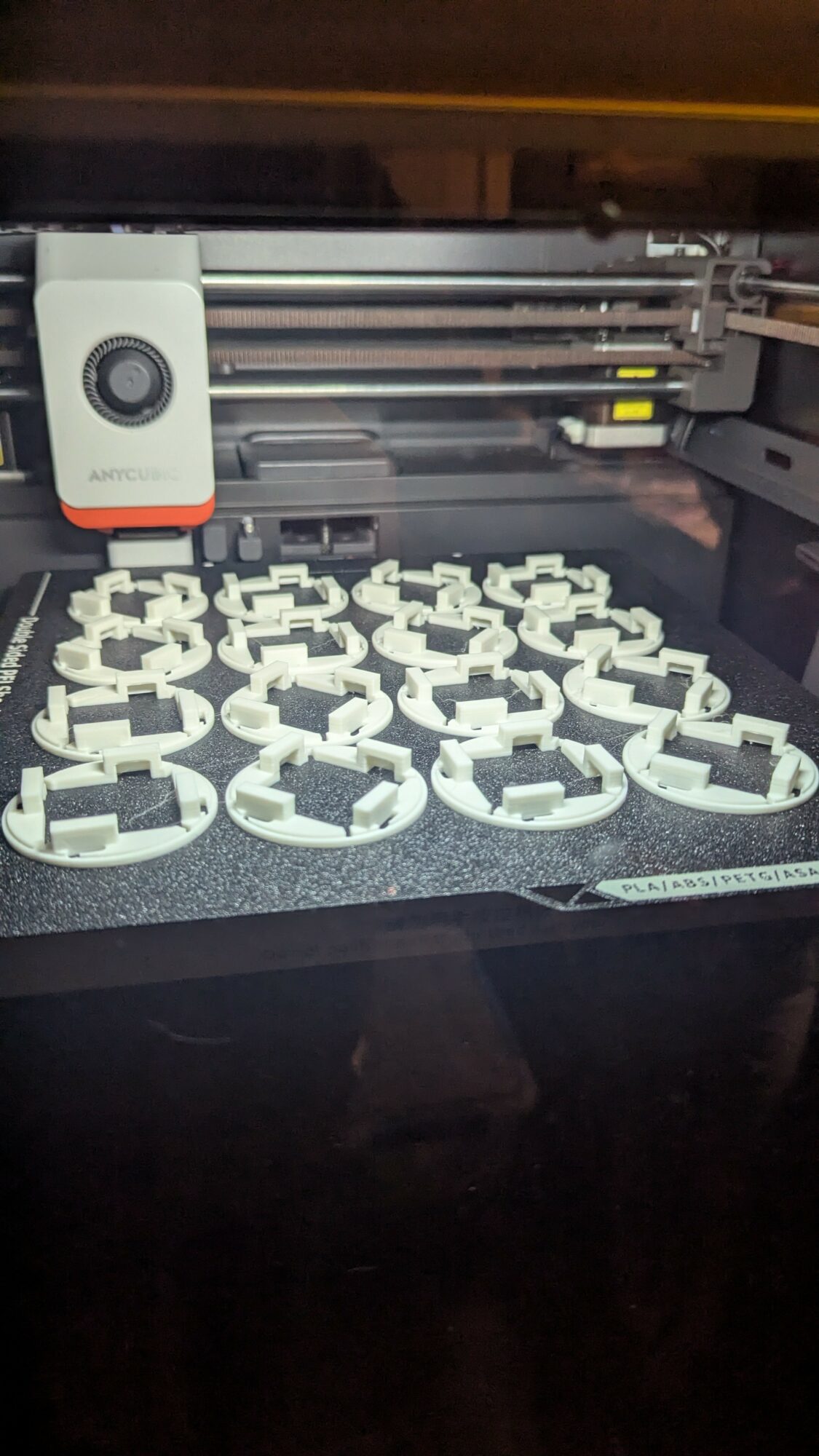 3D printer printing multiple white plastic objects on a black surface, with the printer's extruder and rails visible.