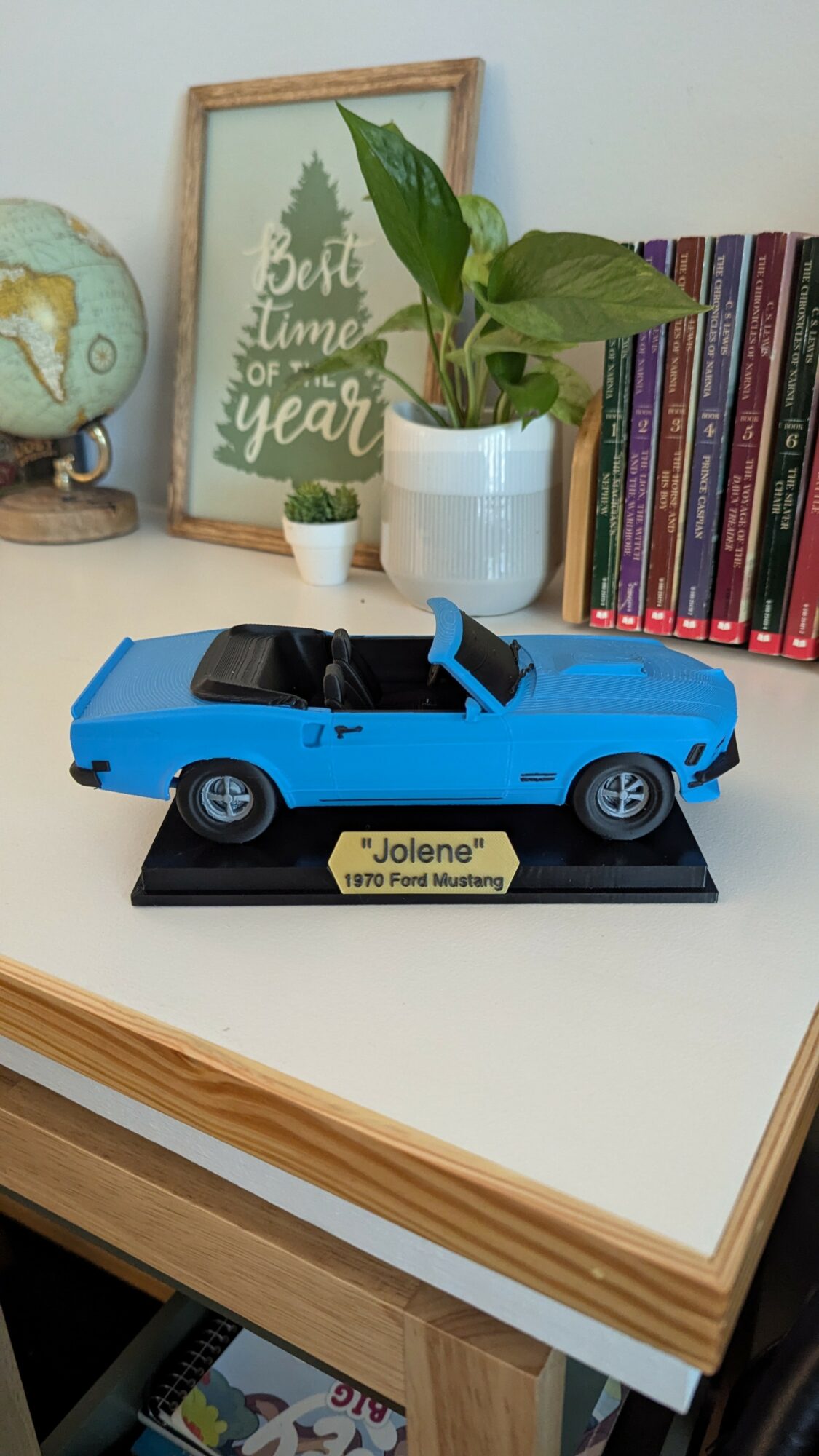 Blue toy car model on a black display base with a yellow nameplate, placed on a white table with books and plants in background.