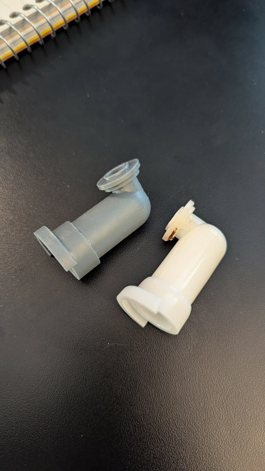 Two plastic connectors on a black surface, one gray and one white, with a spiral notebook partially visible at the top.
