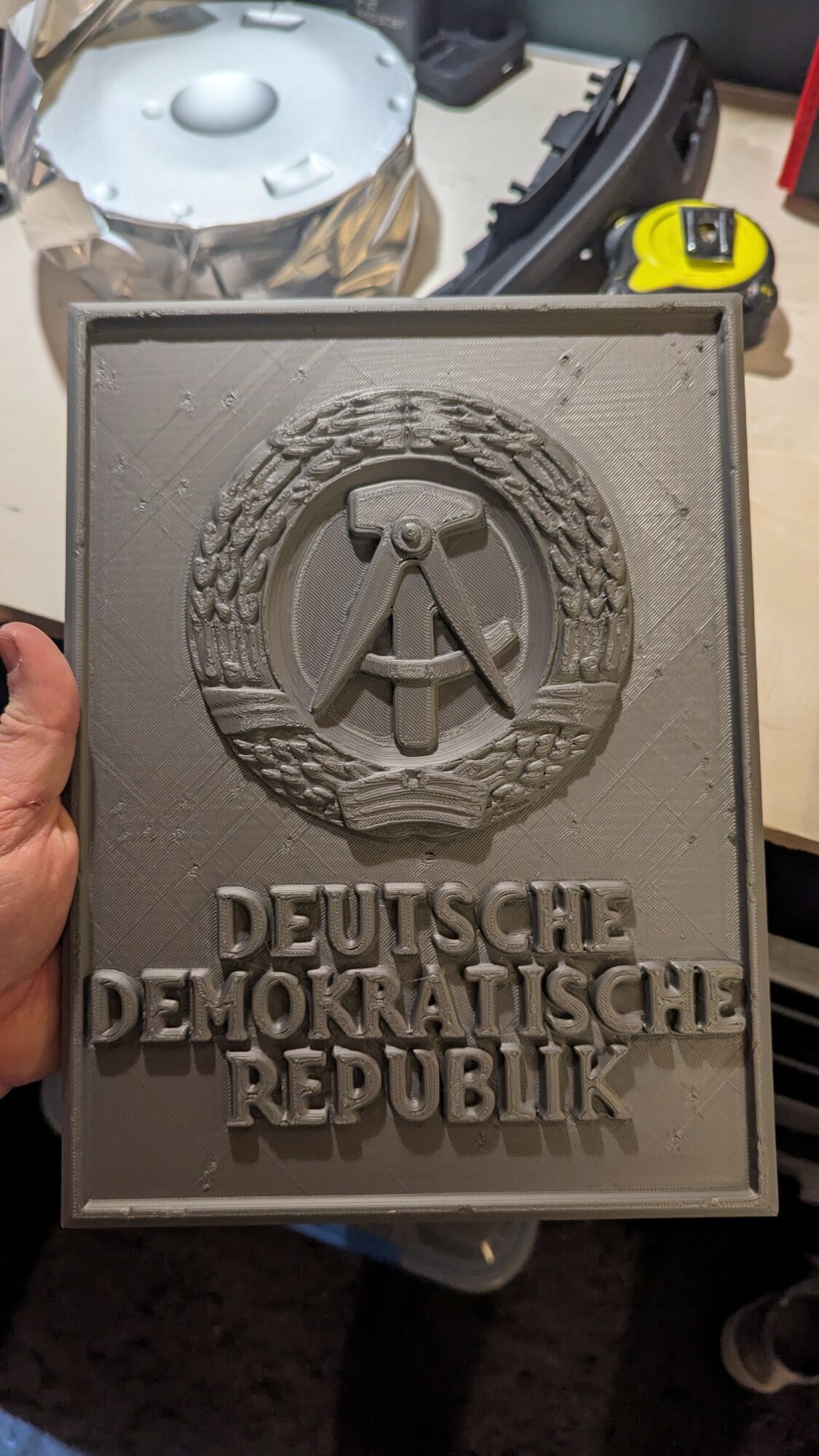 Gray rectangular plaque with emblem and text, held by a hand, background includes a table and tools.