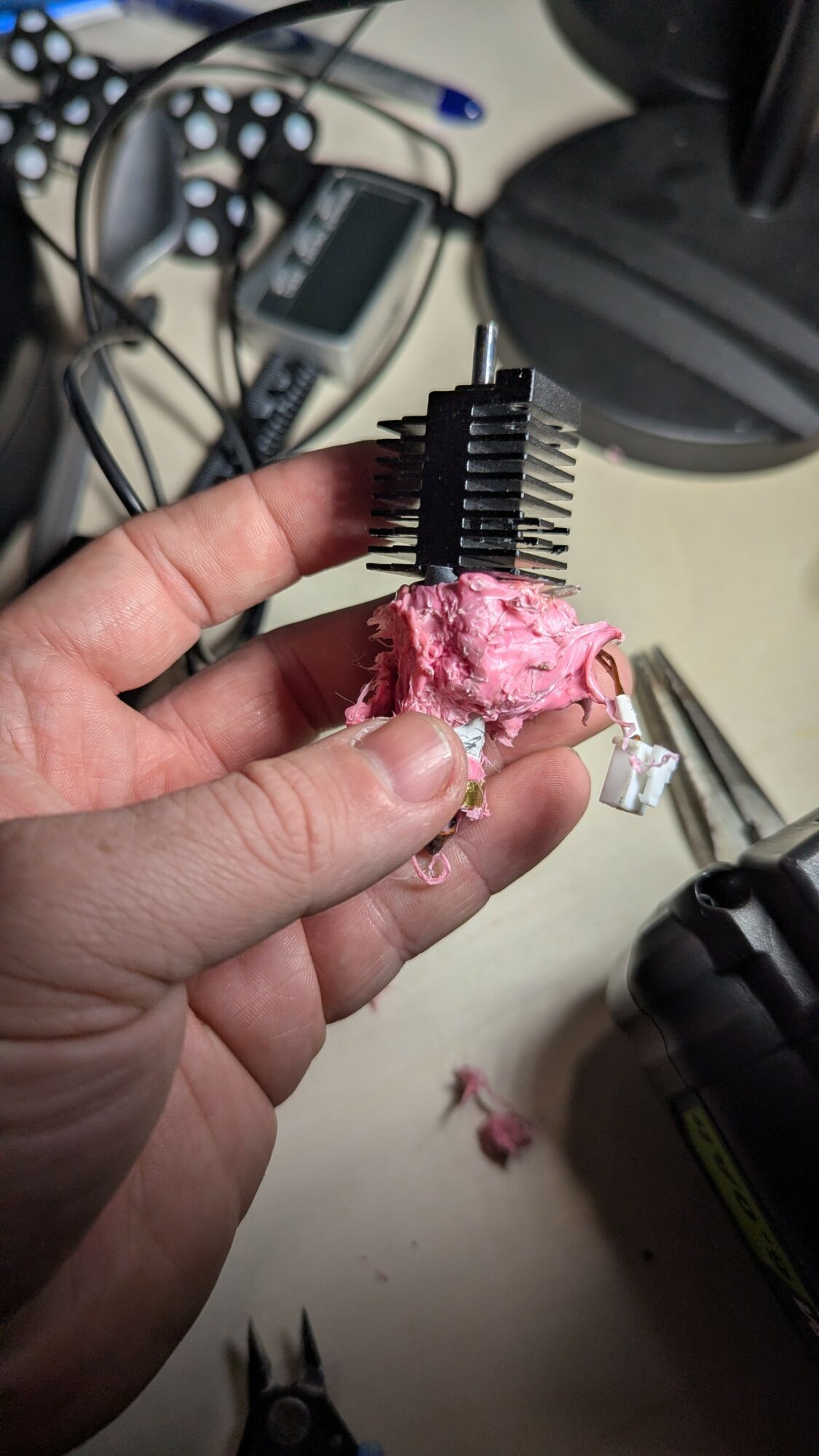 Hand holding a small electronic component with pink material, surrounded by cables and electronic devices on a desk.