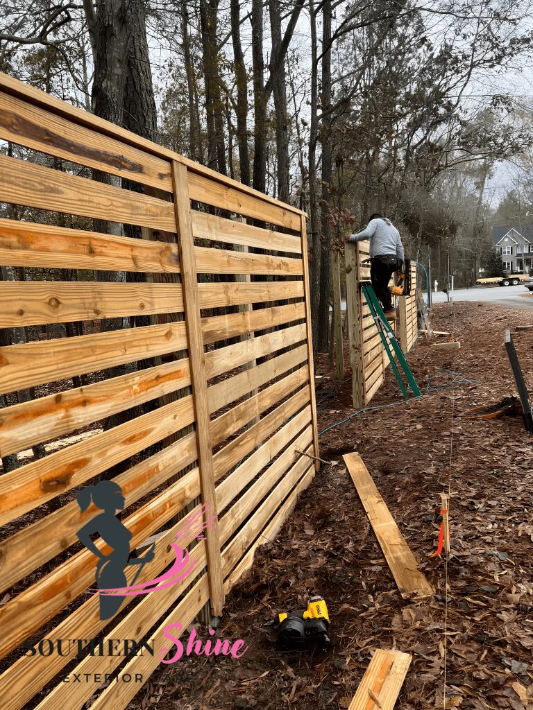 Person working on wooden fence in yard, ladder nearby, trees in background, ground covered with soil and wood planks.