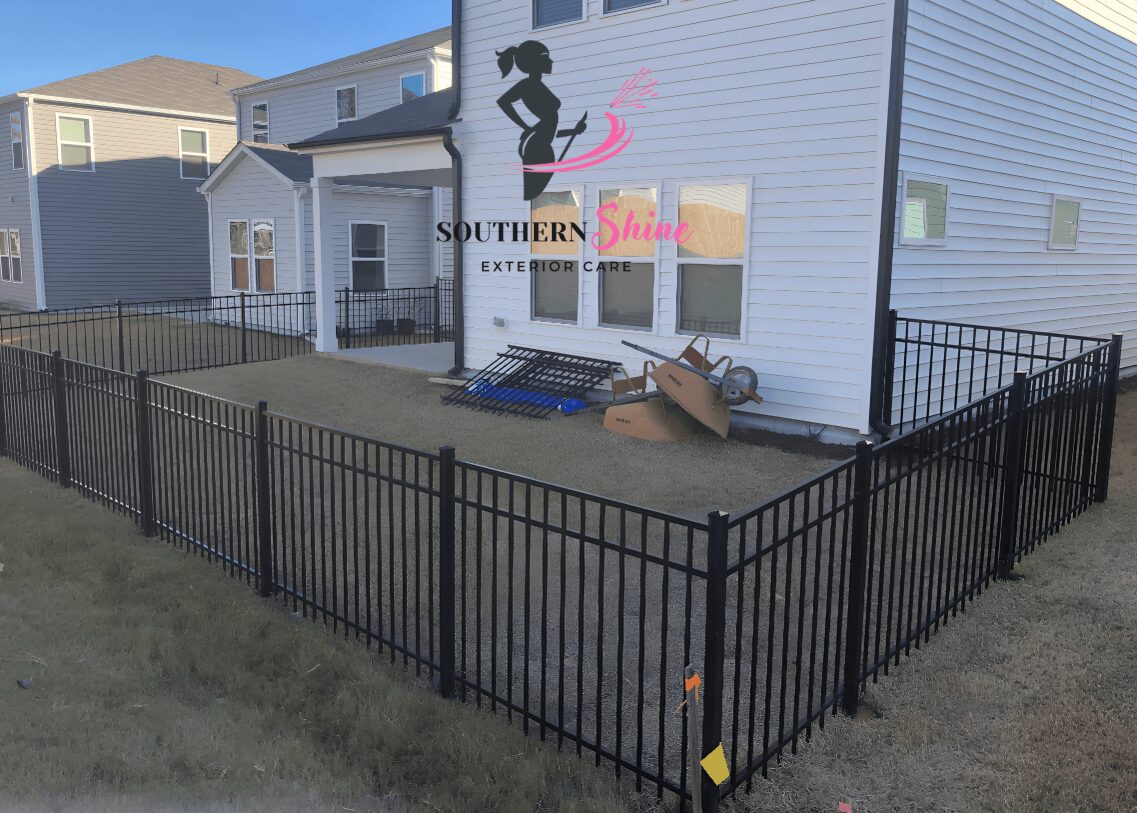 Fenced outdoor area with a bench and some boxes near a white house with windows.