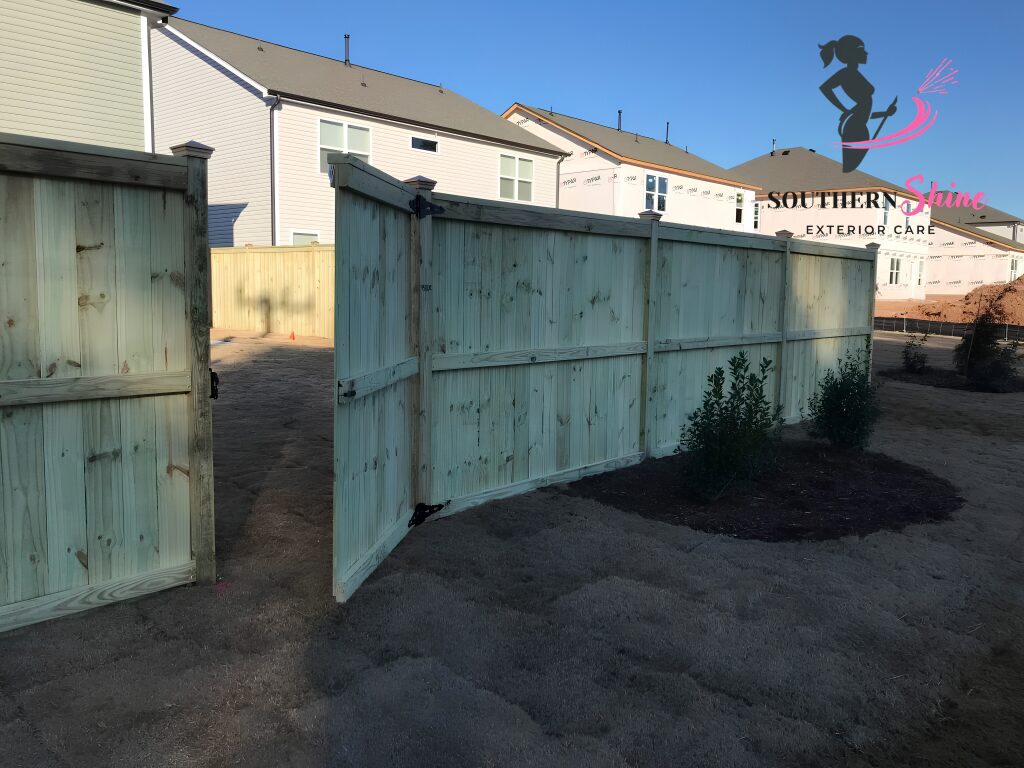 Wooden fence with a small bush and dirt ground in front, residential buildings in background under blue sky.