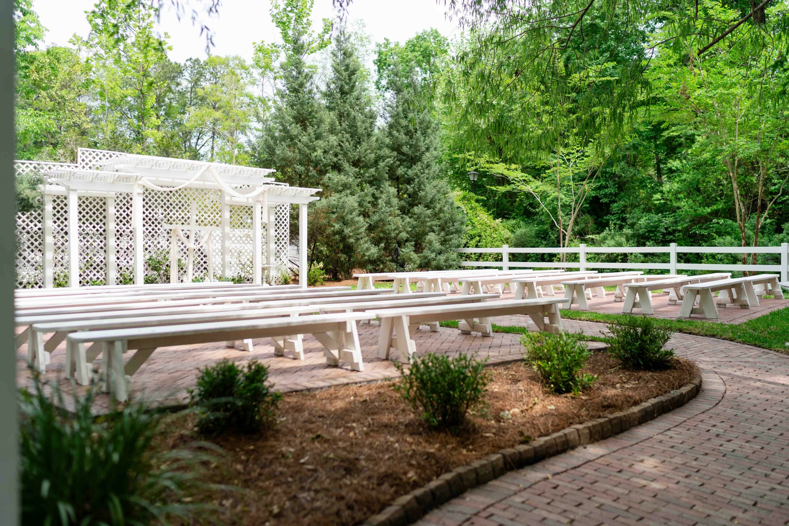 Outdoor seating area with white benches, surrounded by trees and greenery, paved walkway, and small bushes.