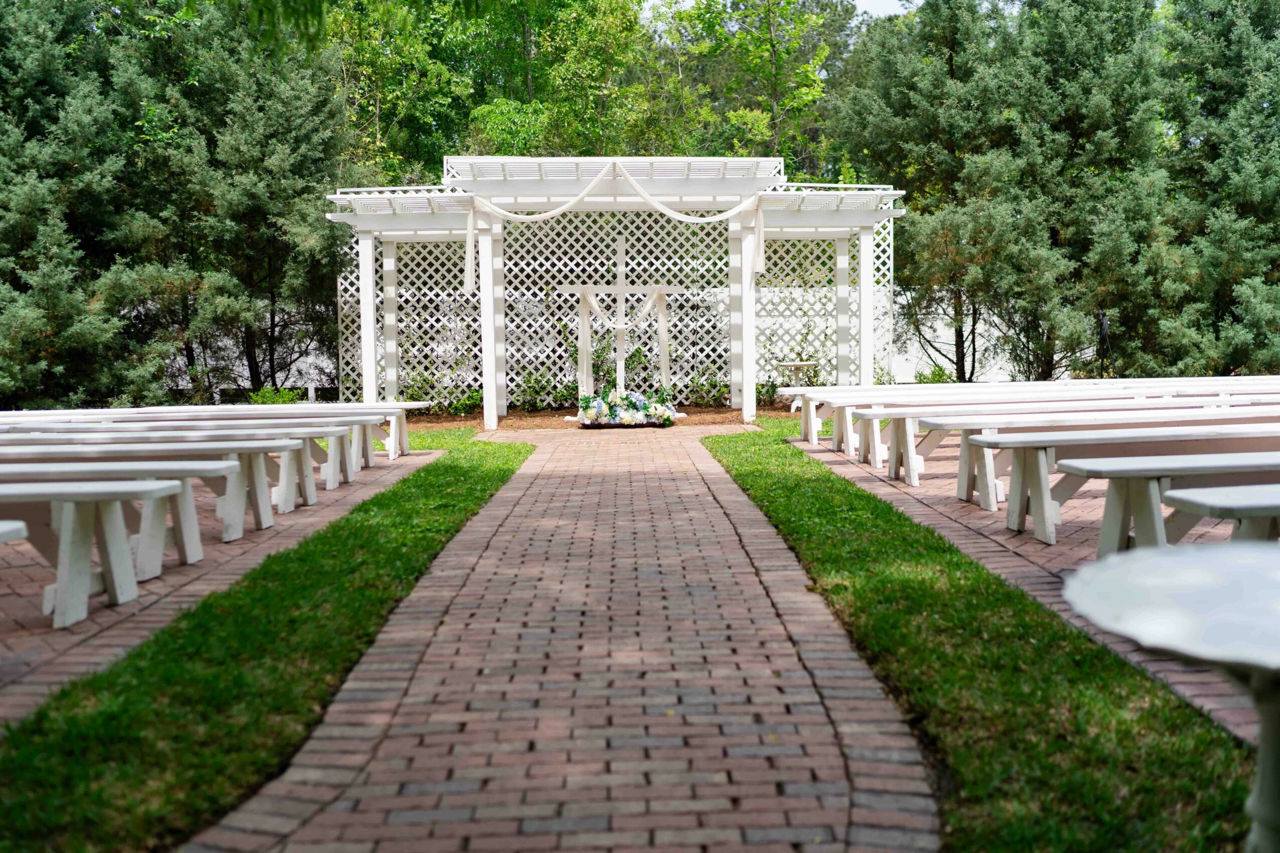 Outdoor wedding venue with white pergola, brick pathway, benches on both sides, surrounded by green trees.