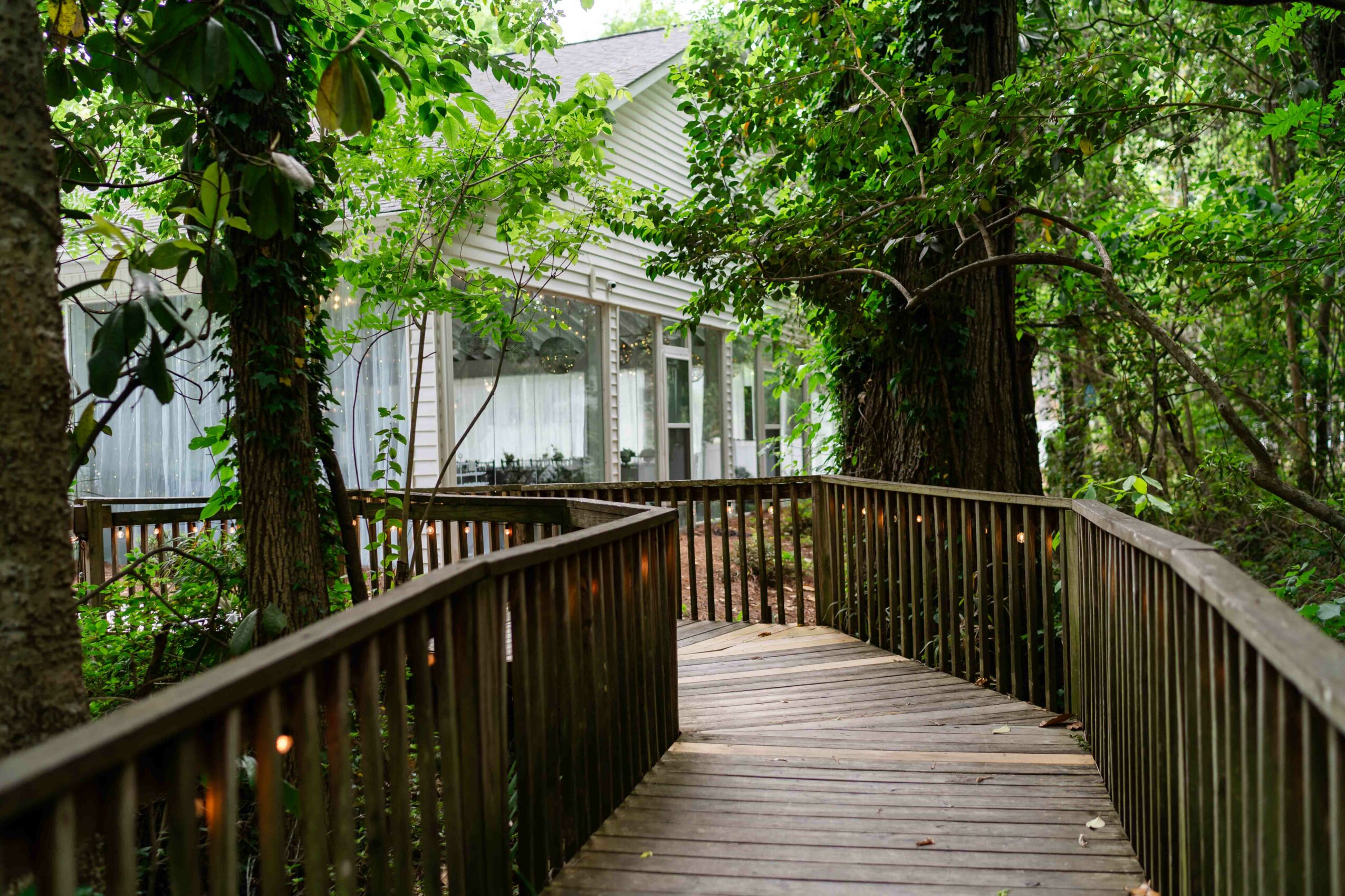 Wooden pathway with railings curves through a green wooded area near a house with large windows.