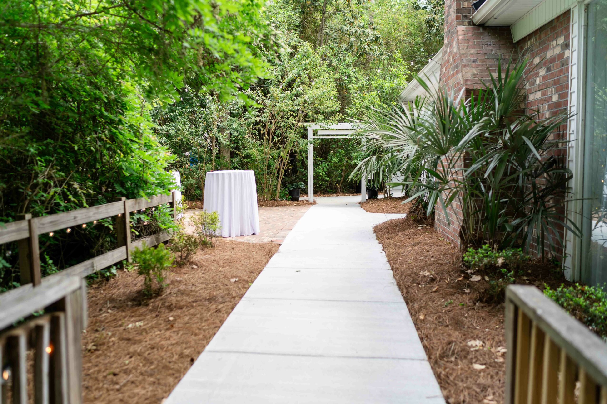 Concrete pathway leading through a garden area with plants and trees, bordered by a wooden fence and a brick building.